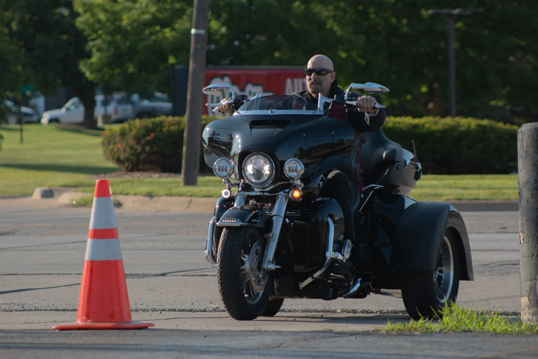 Bald man wearing sunglasses riding a black motorcycle through an area with orange traffic cones, in a park-like setting with trees and cars in the background.