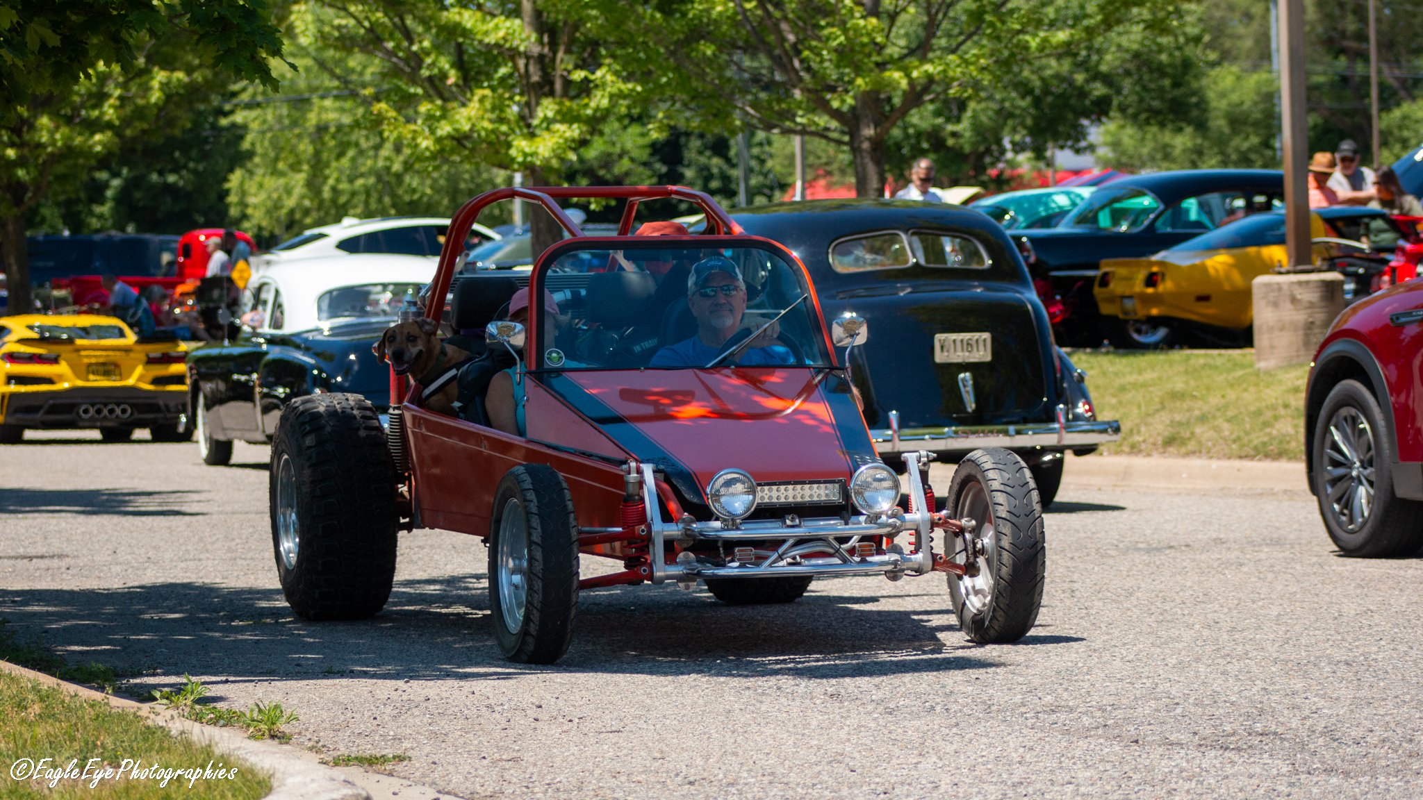 Red off-road vehicle with a black roll cage and large tires driving through a car show, surrounded by classic and sports cars under trees.