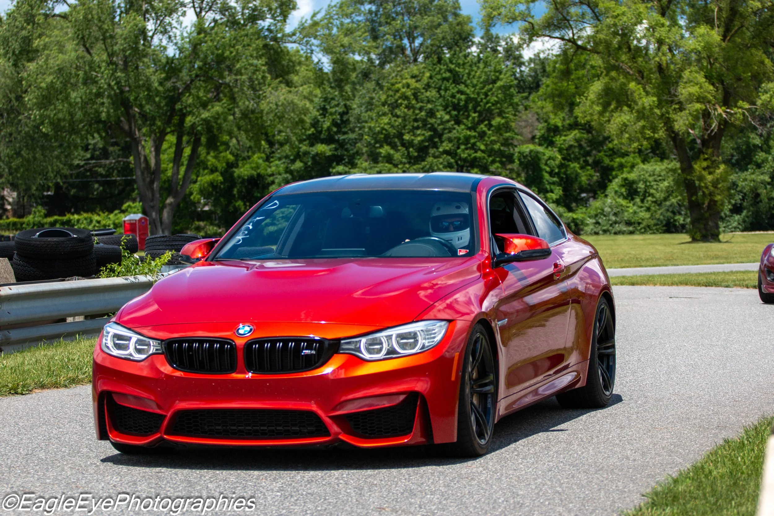 A red sports car with a black roof and frontal LED lights, parked on a street with lush green trees in the background.