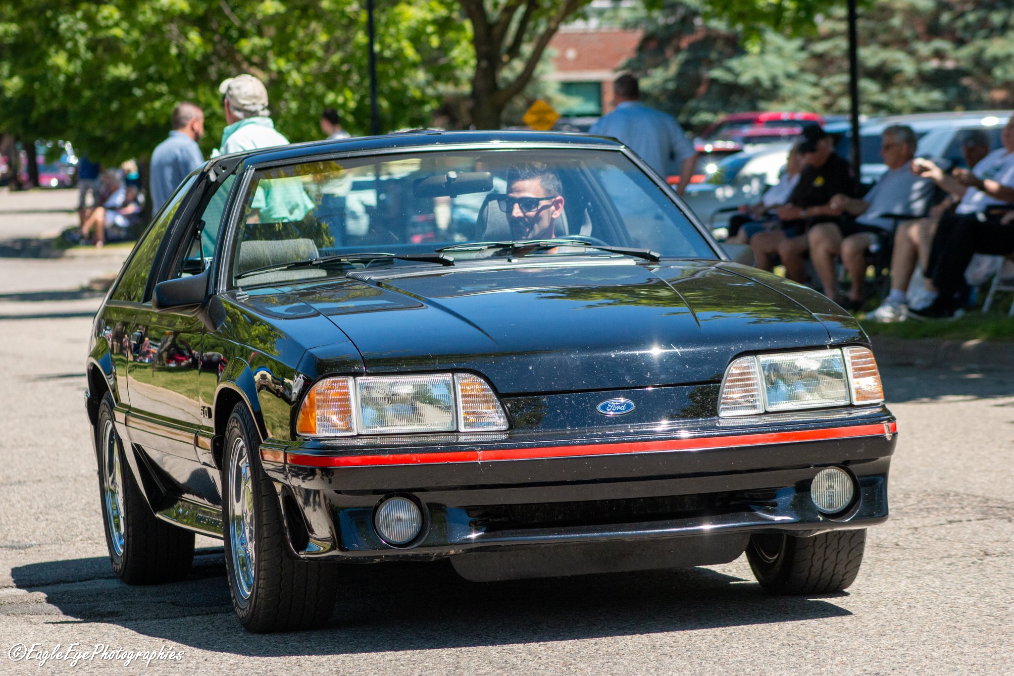 A black vintage Ford Mustang convertible car with a male driver wearing sunglasses, parked on a street with people sitting on benches and standing around, lush green trees, and buildings in the background.