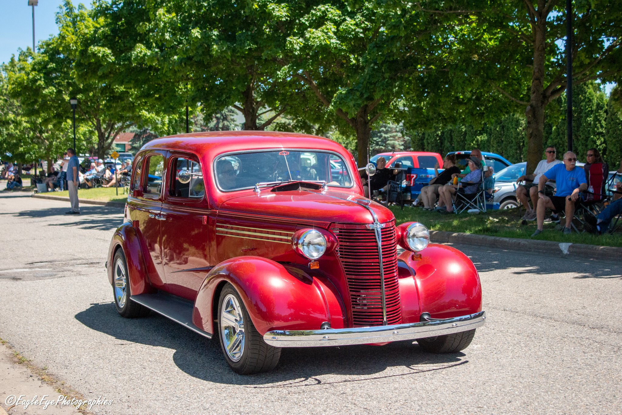 Red vintage car at a car show with people sitting on lawn chairs and a large green leafy tree in the background.