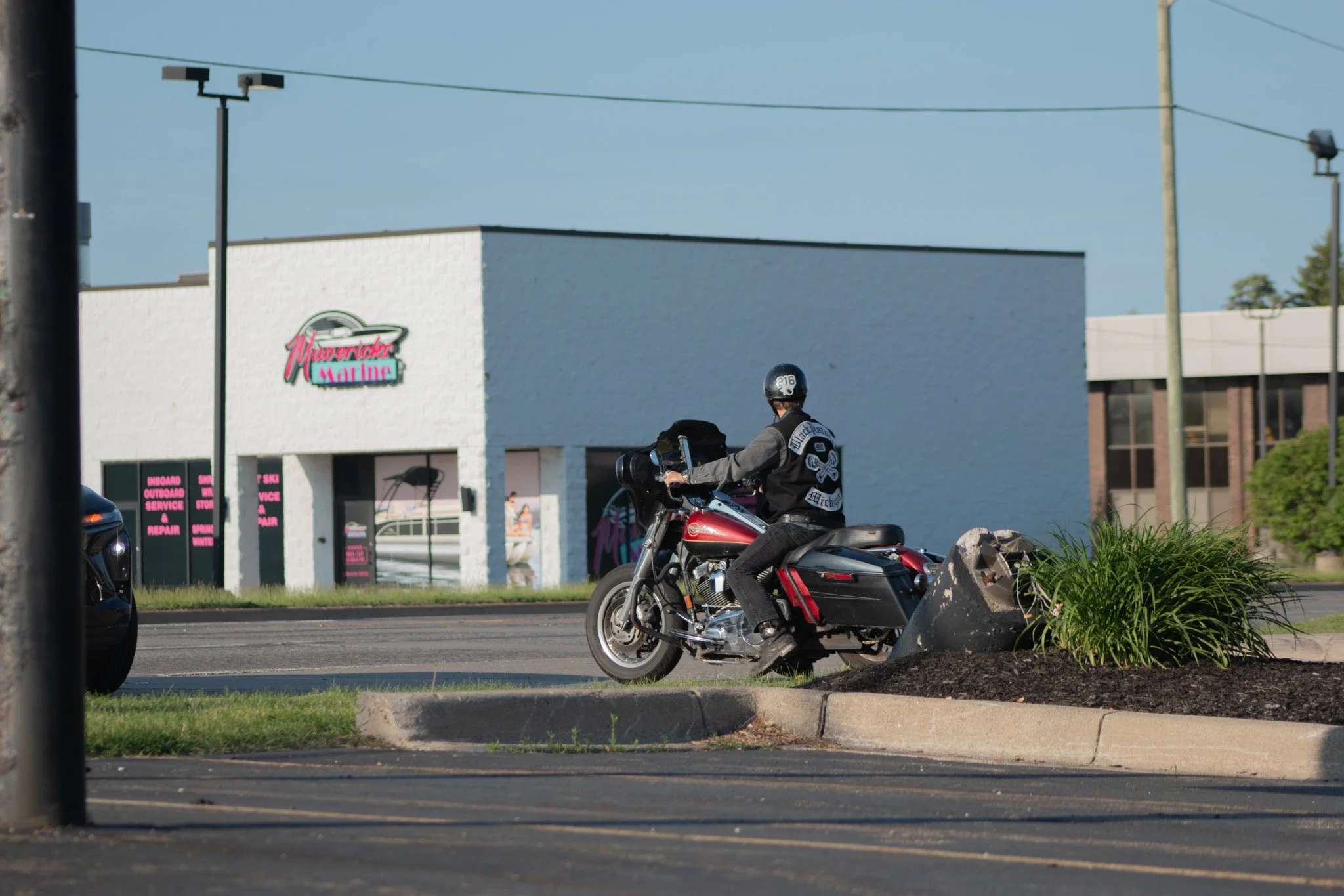 A person riding a red motorcycle wearing a black helmet and jacket in front of a white building with a pink and neon sign that says 'Maranda's Marine.'