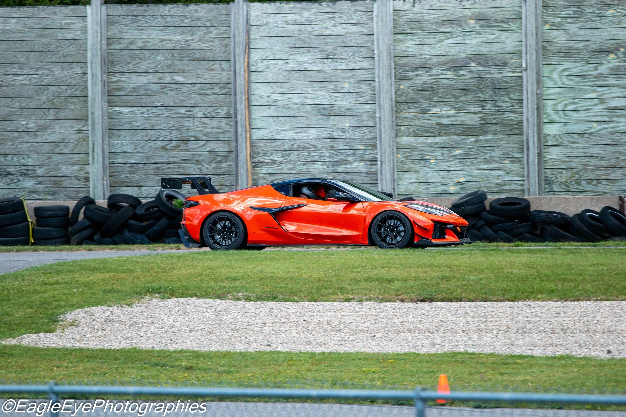 A bright orange sports car parked on a grass area beside a paved road, with black racing tires stacked along a concrete barrier and a wooden fence in the background.