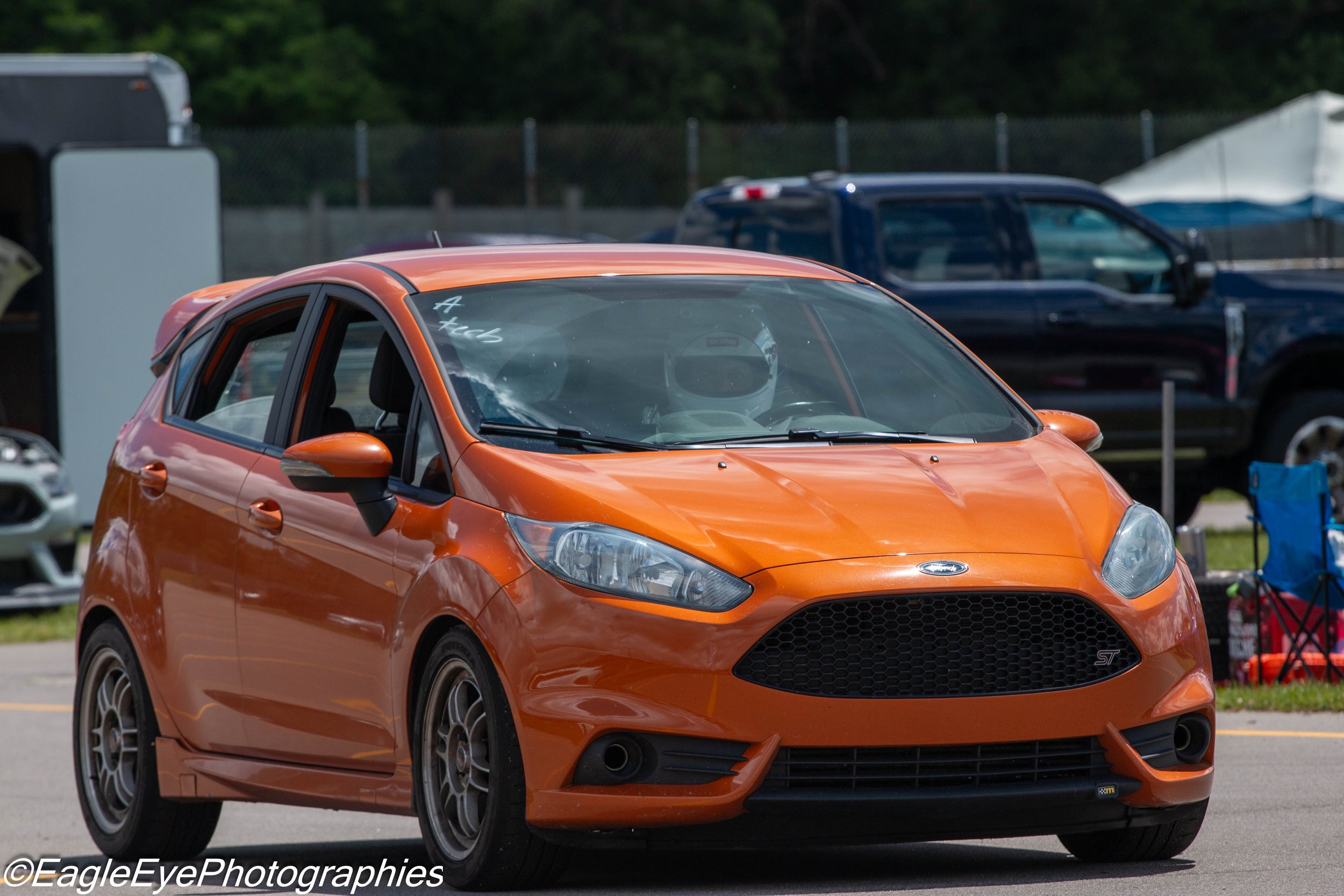 An orange Ford Fiesta parked in a parking lot with other vehicles and tents in the background.