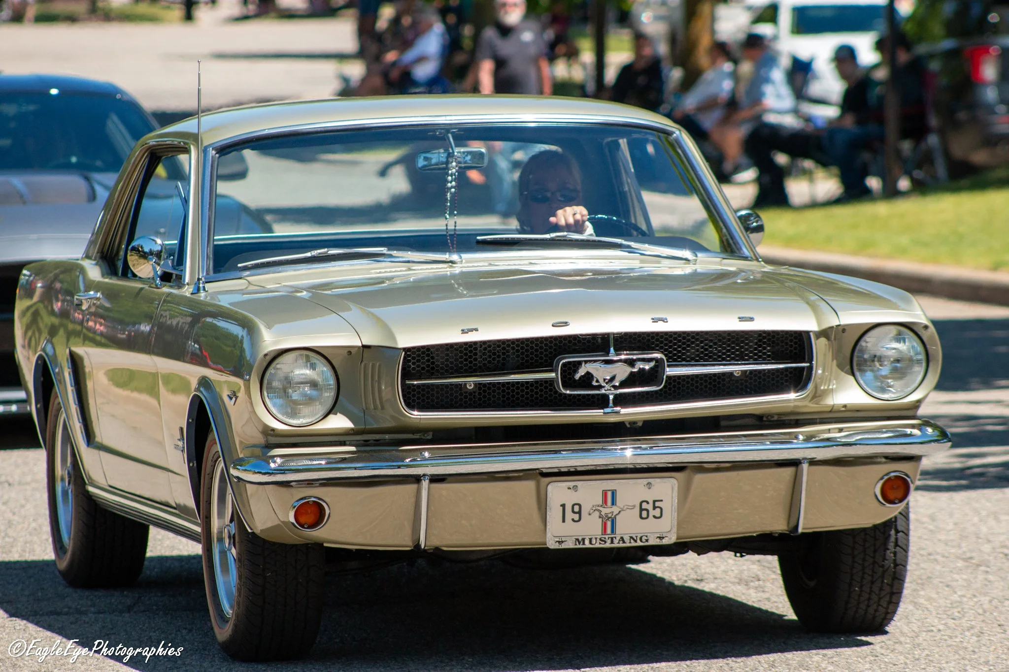 A vintage golden 1965 Ford Mustang parked outdoors during a car show, with people in the background.