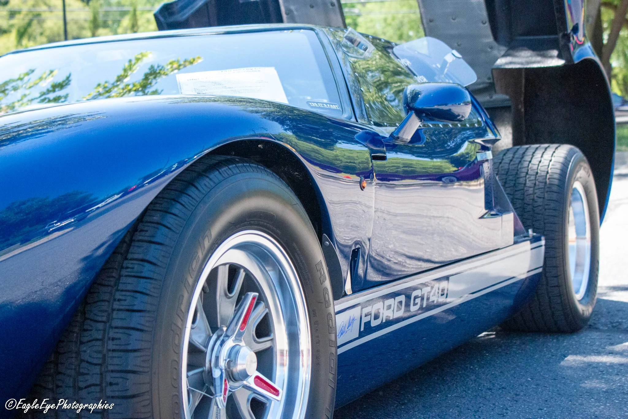 Close-up of a blue Ford GT40 race car with visible tire and wheel, windshield with document, and side mirror, parked outdoors with trees reflected on its surface.