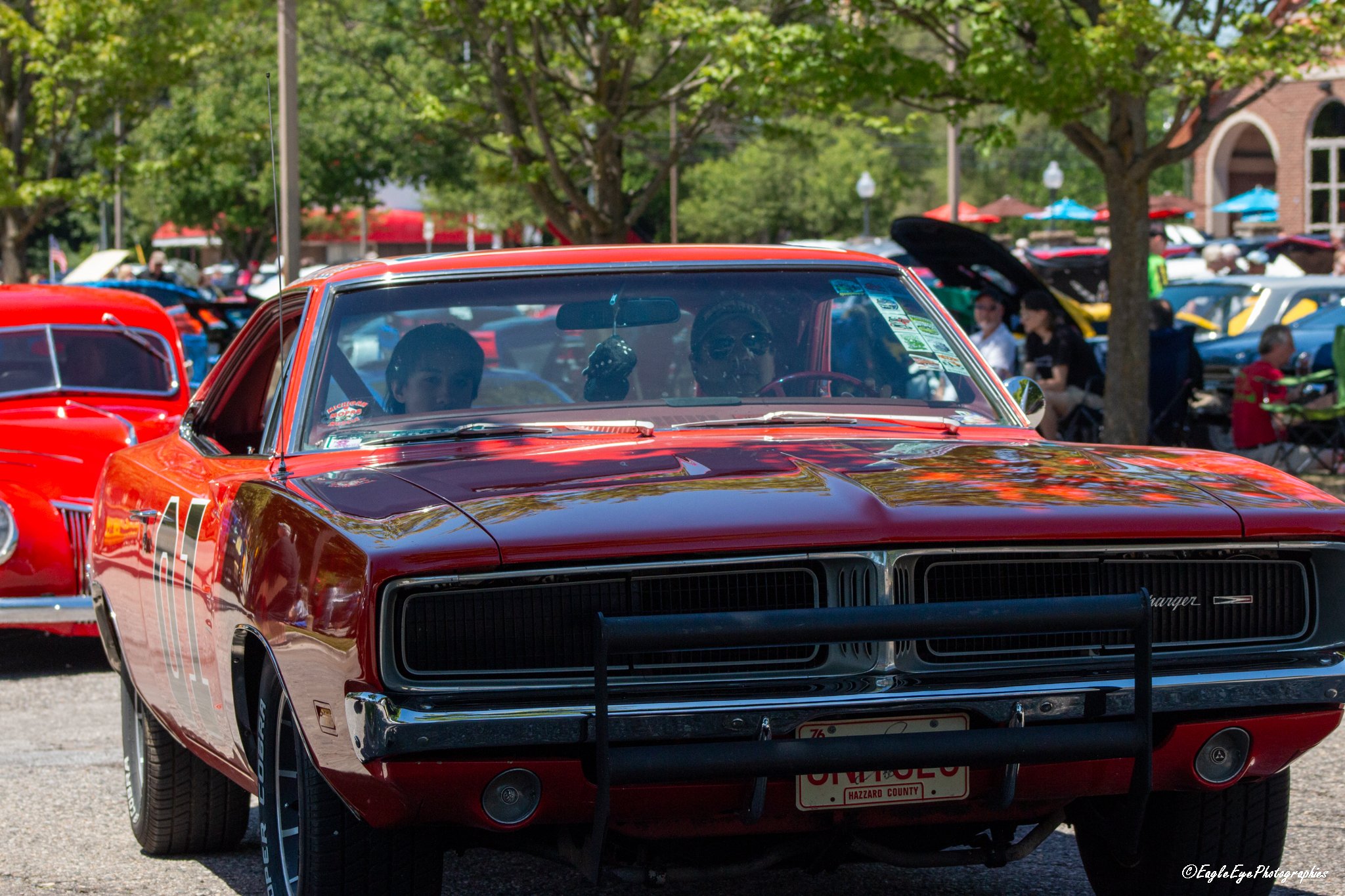 A red vintage muscle car with black grille and chrome details at an outdoor car show, with people and other classic cars in the background under trees.