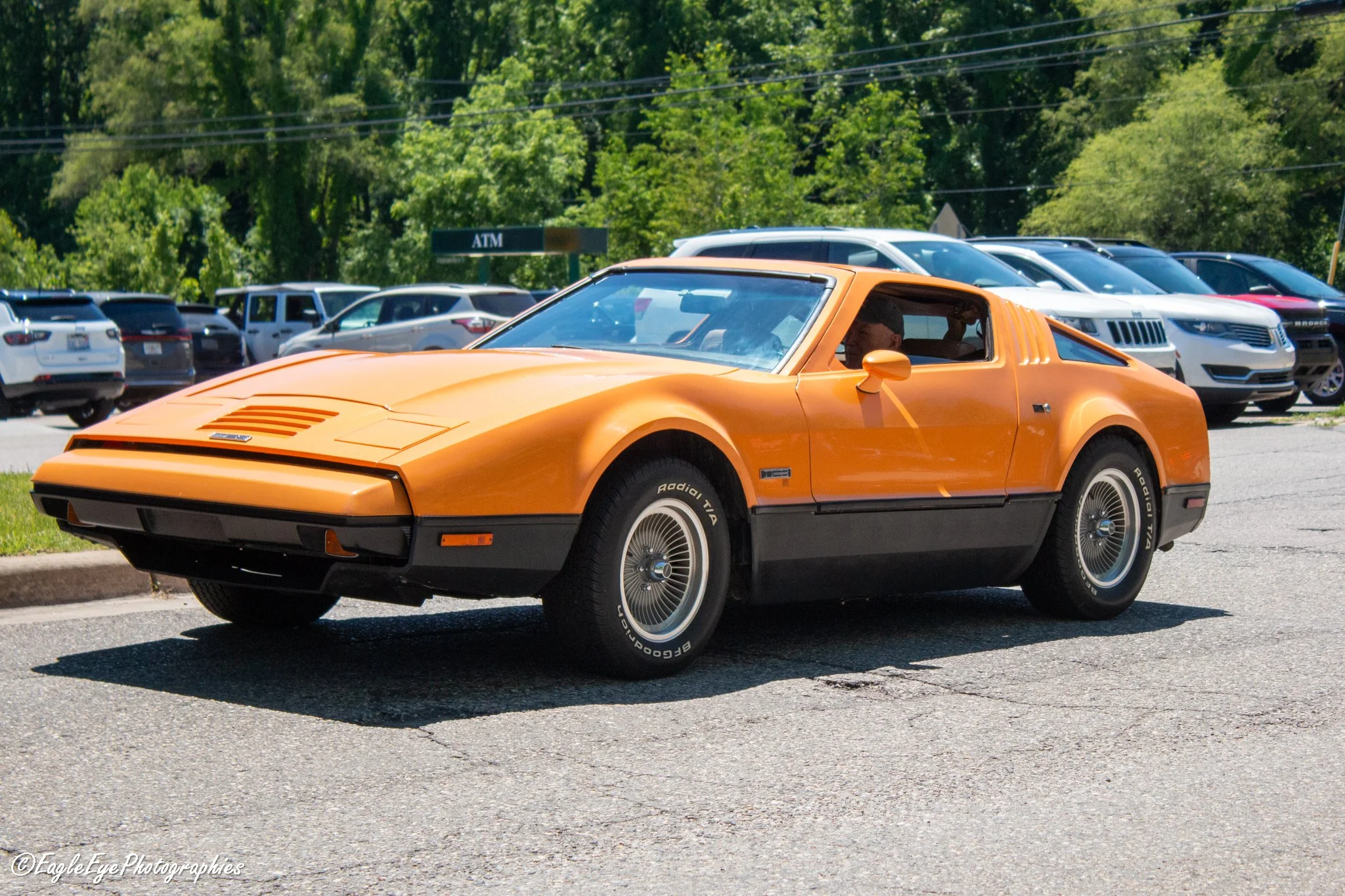 An orange vintage sports car parked in a parking lot with other modern vehicles, trees in the background, and a sign indicating an ATM.