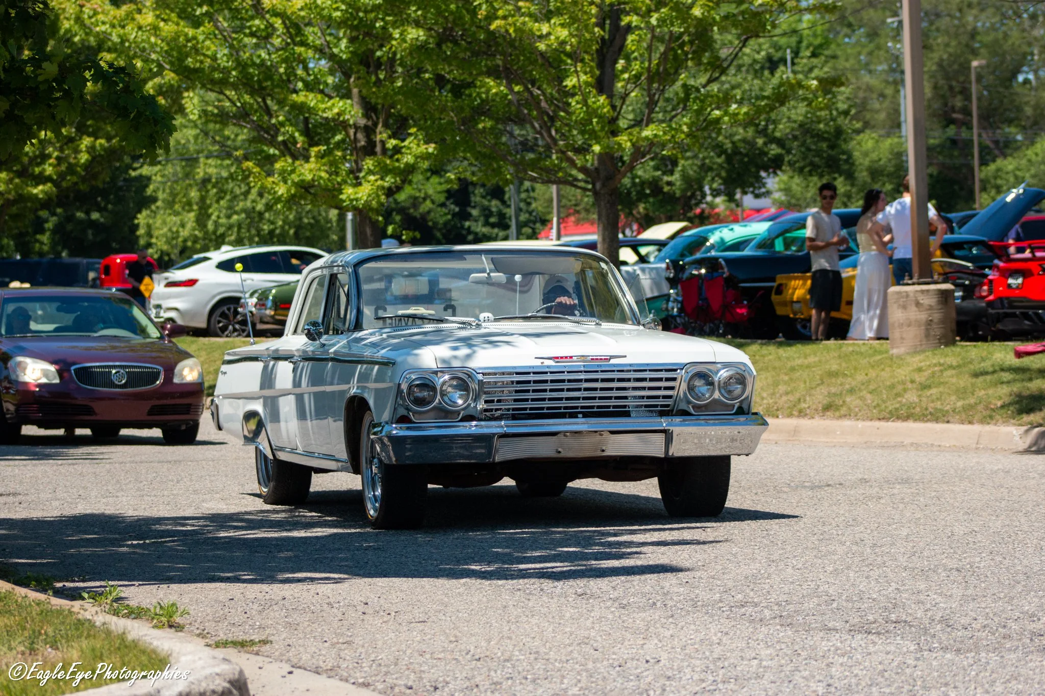 A vintage white car parked in a parking lot during daytime with several modern cars and people in the background, underneath trees with green leaves.