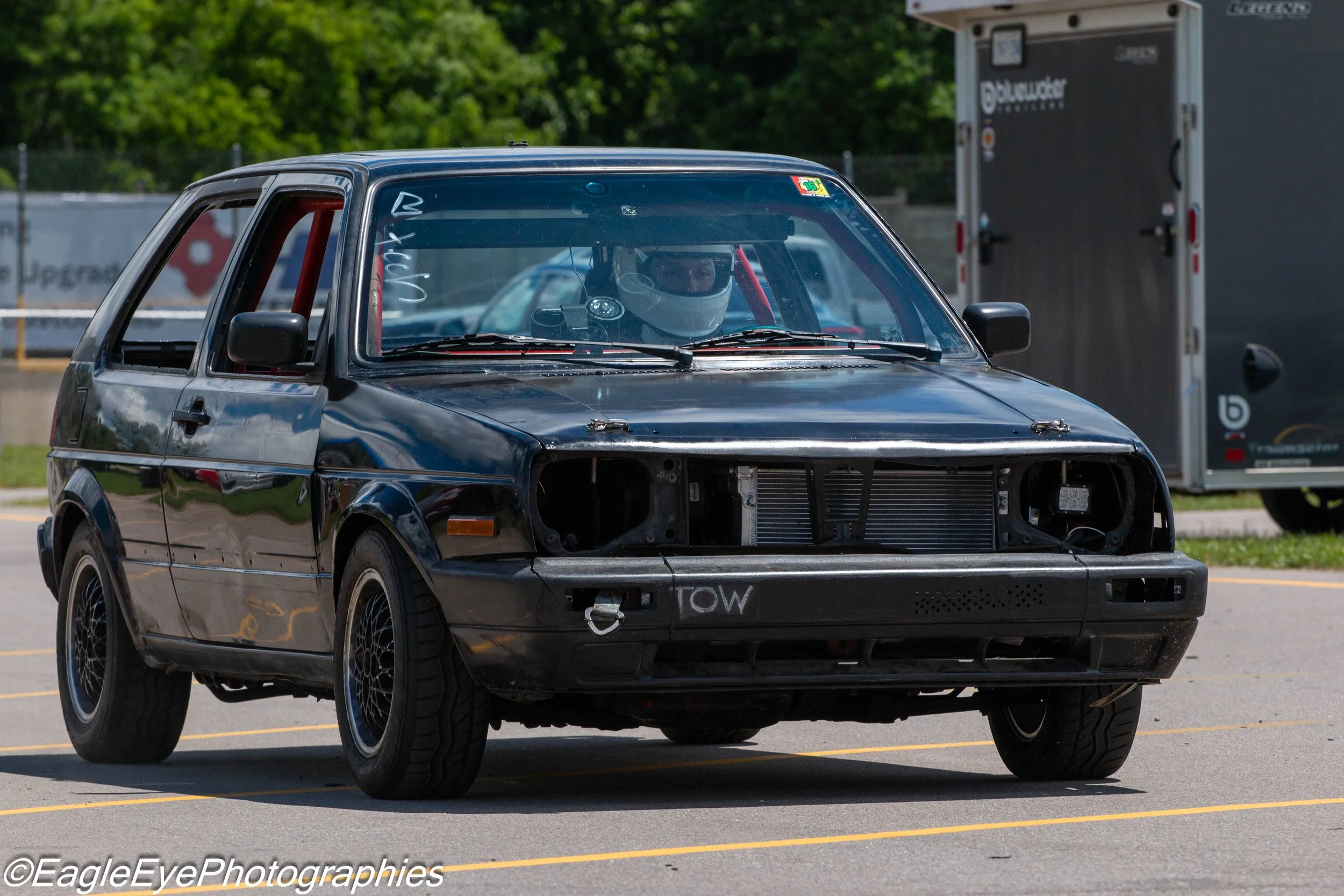 A black race car with no front bumper revealing the radiator and engine components, driver wearing a white helmet, in a parking lot with trailer and trees in the background.