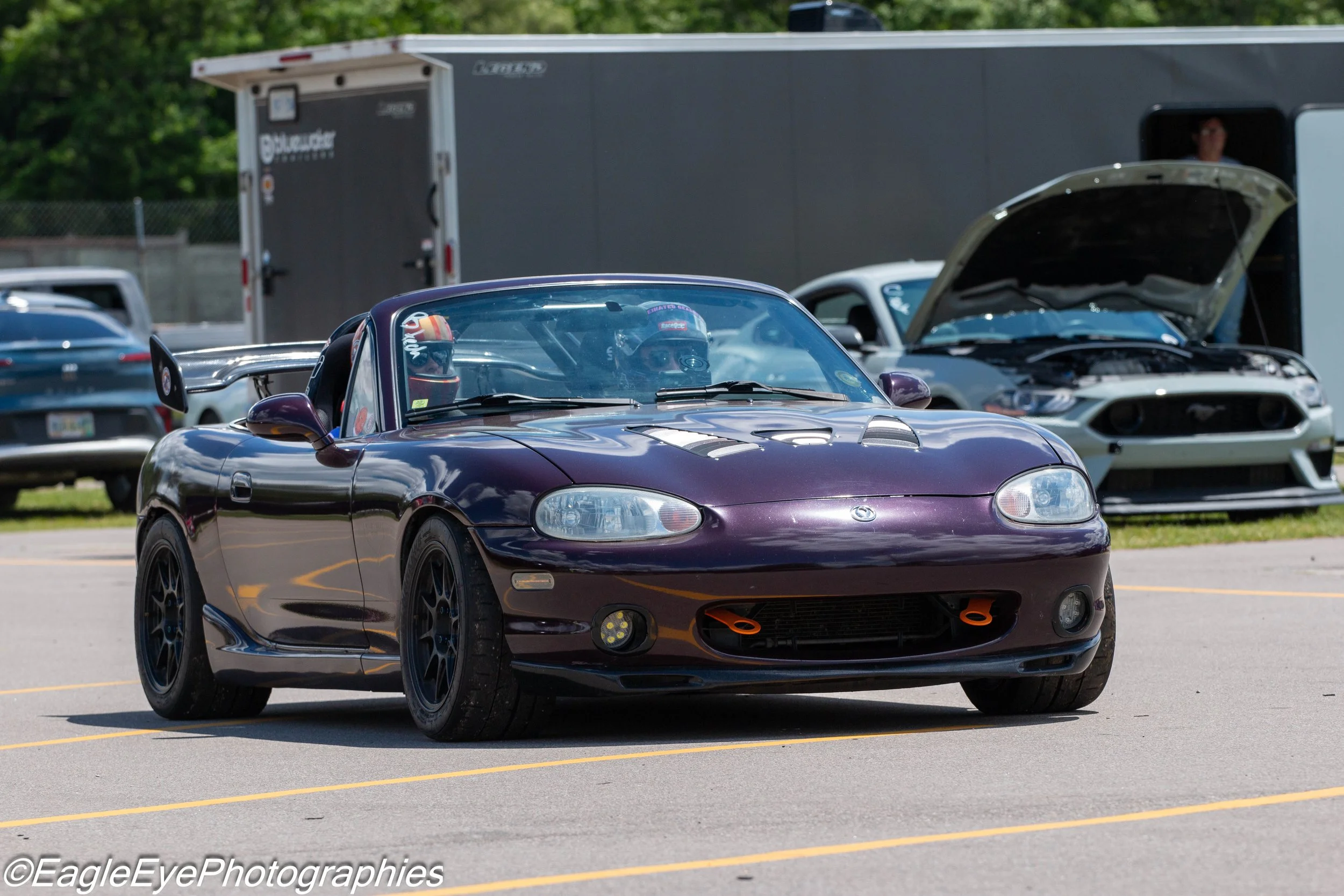 Purple Mazda Miata sports car with a driver and passenger in racing helmets parked in a lot, with other cars and a trailer in the background.