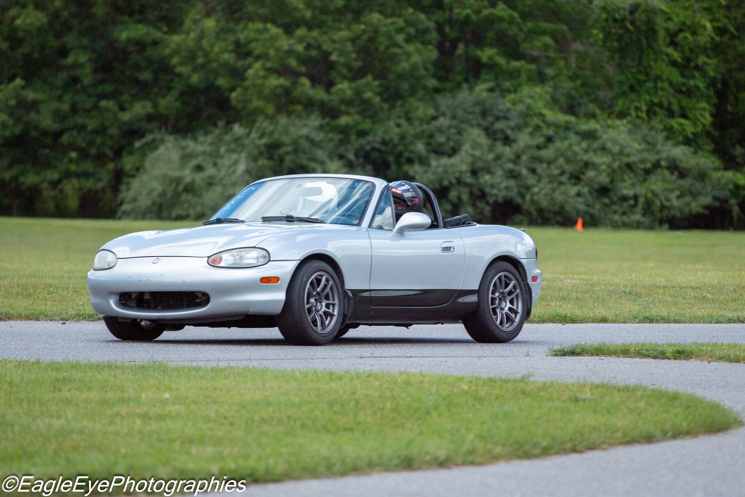 A silver Mazda MX-5 convertible driving on a grassy field with trees in the background.