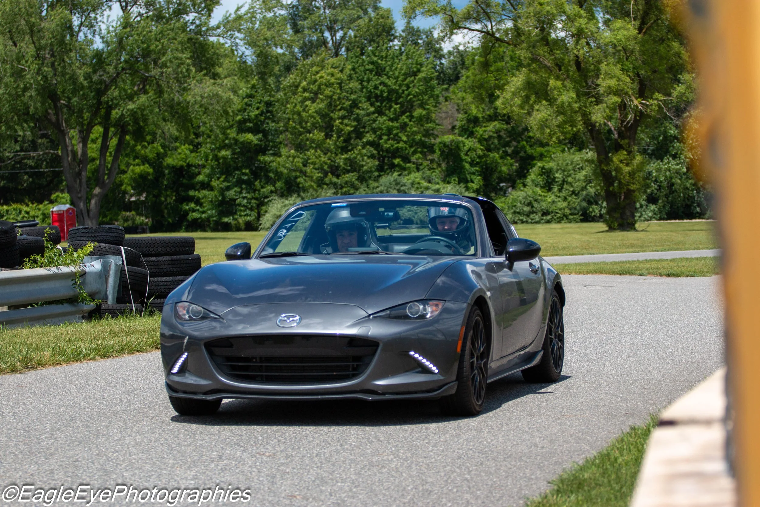 A gray Mazda sports car on a race track, with two drivers wearing helmets inside. The background features green trees and a stack of tires on the side of the track.