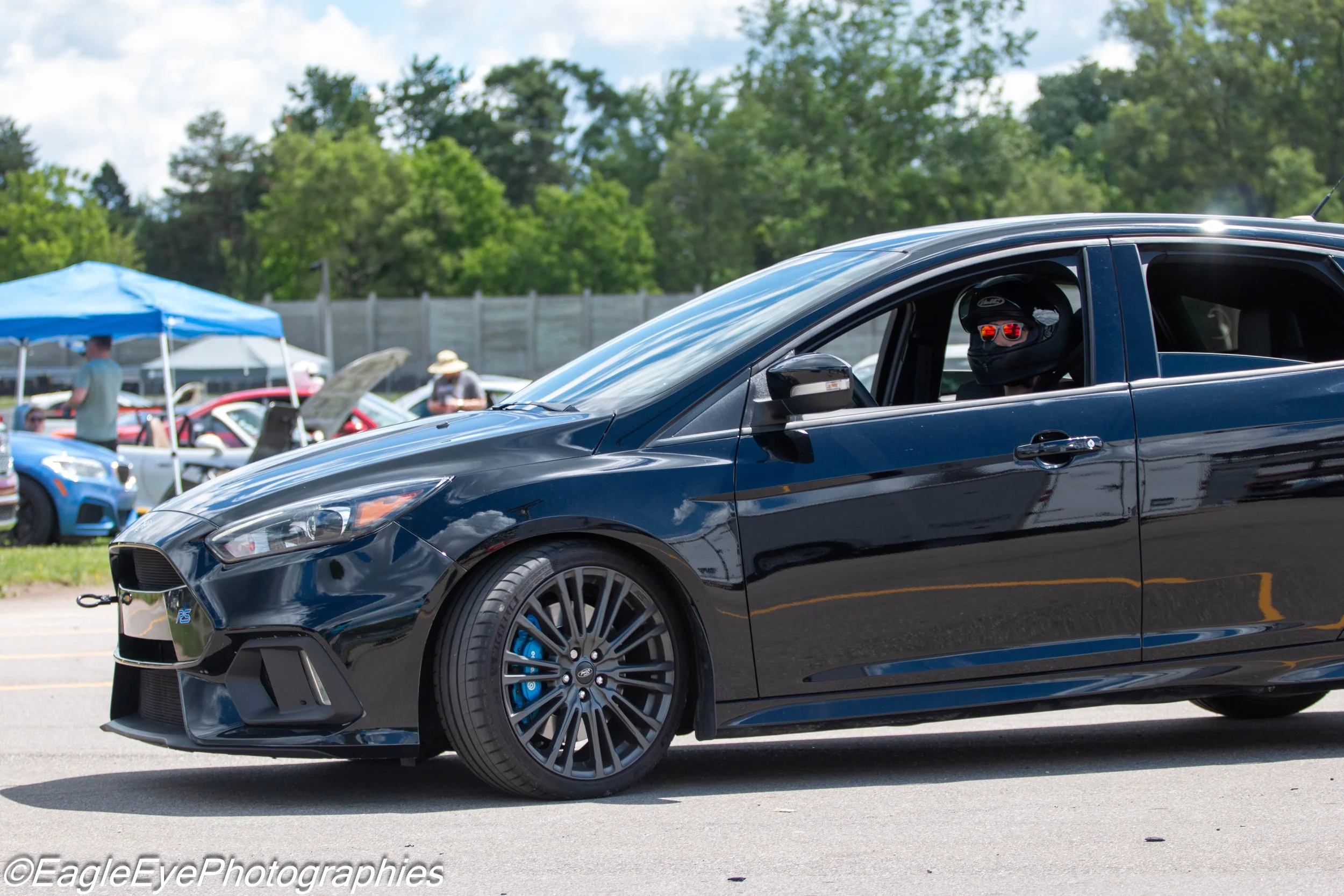 A black sports car with a person wearing a helmet and sunglasses inside, parked at a car show with other cars and tents in the background.