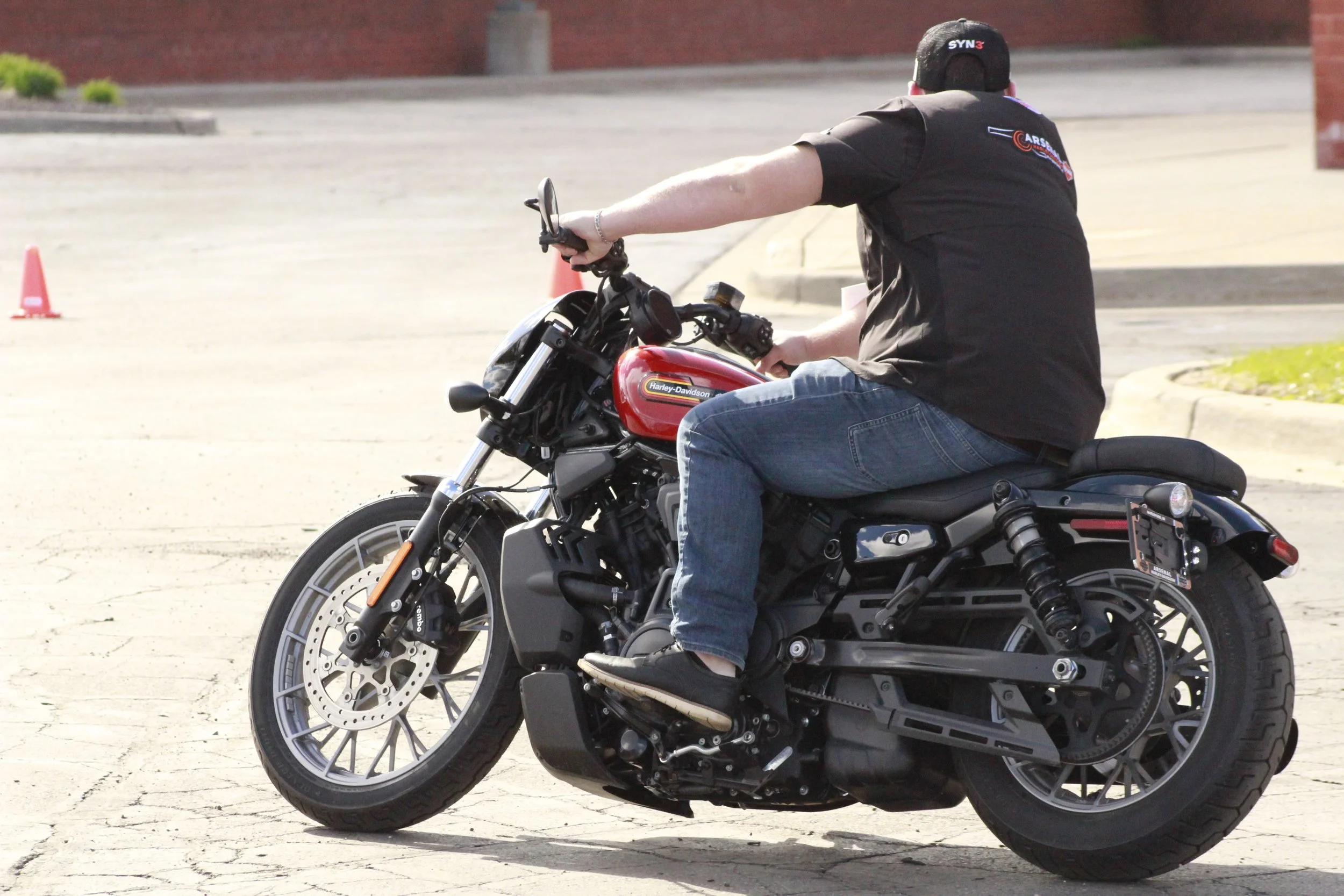 A man riding a black and red Harley-Davidson motorcycle on a paved surface, with orange traffic cones in the background.