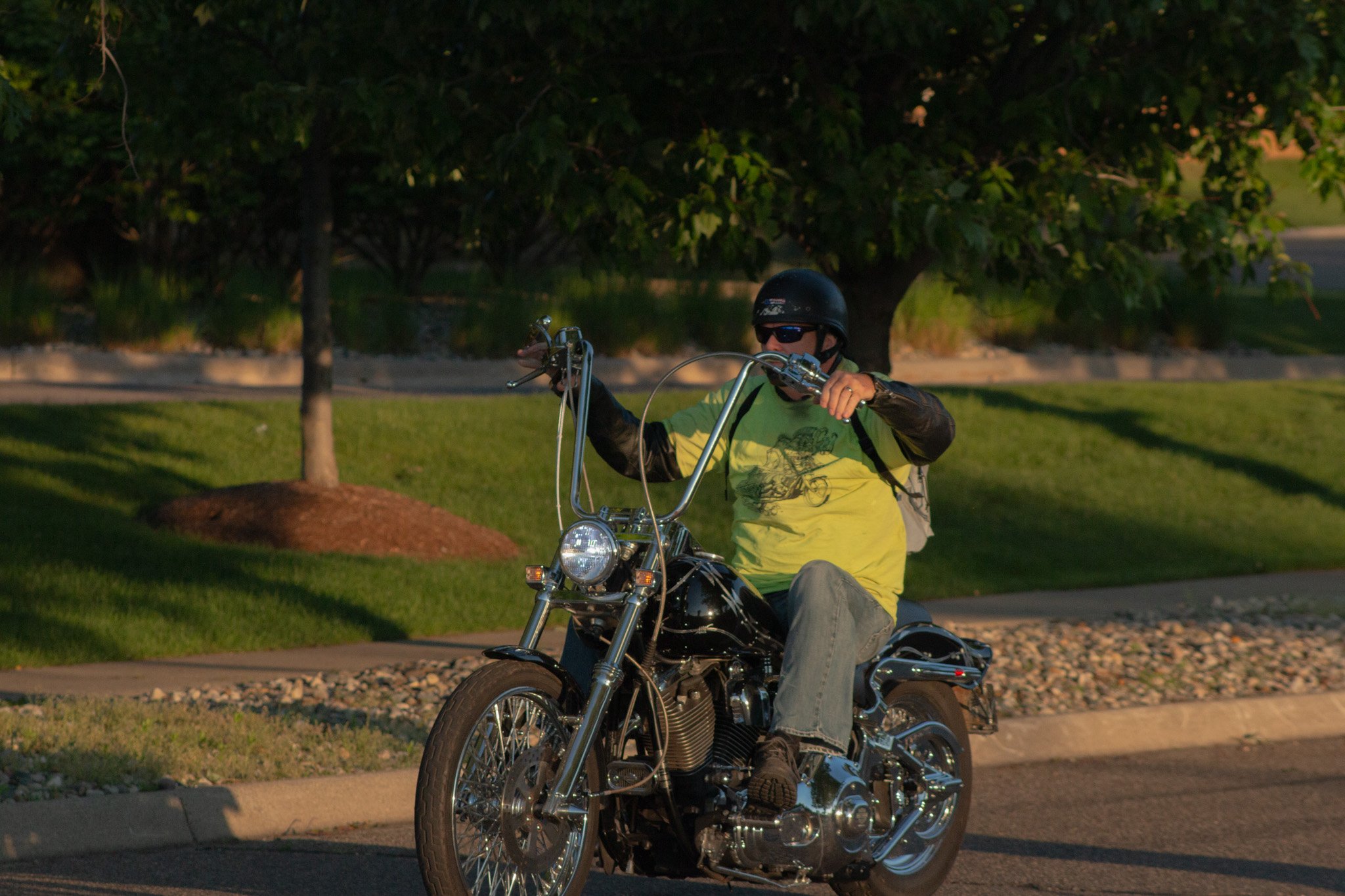 Person wearing a helmet and sunglasses riding a black motorcycle on a road, surrounded by trees and greenery in the background.