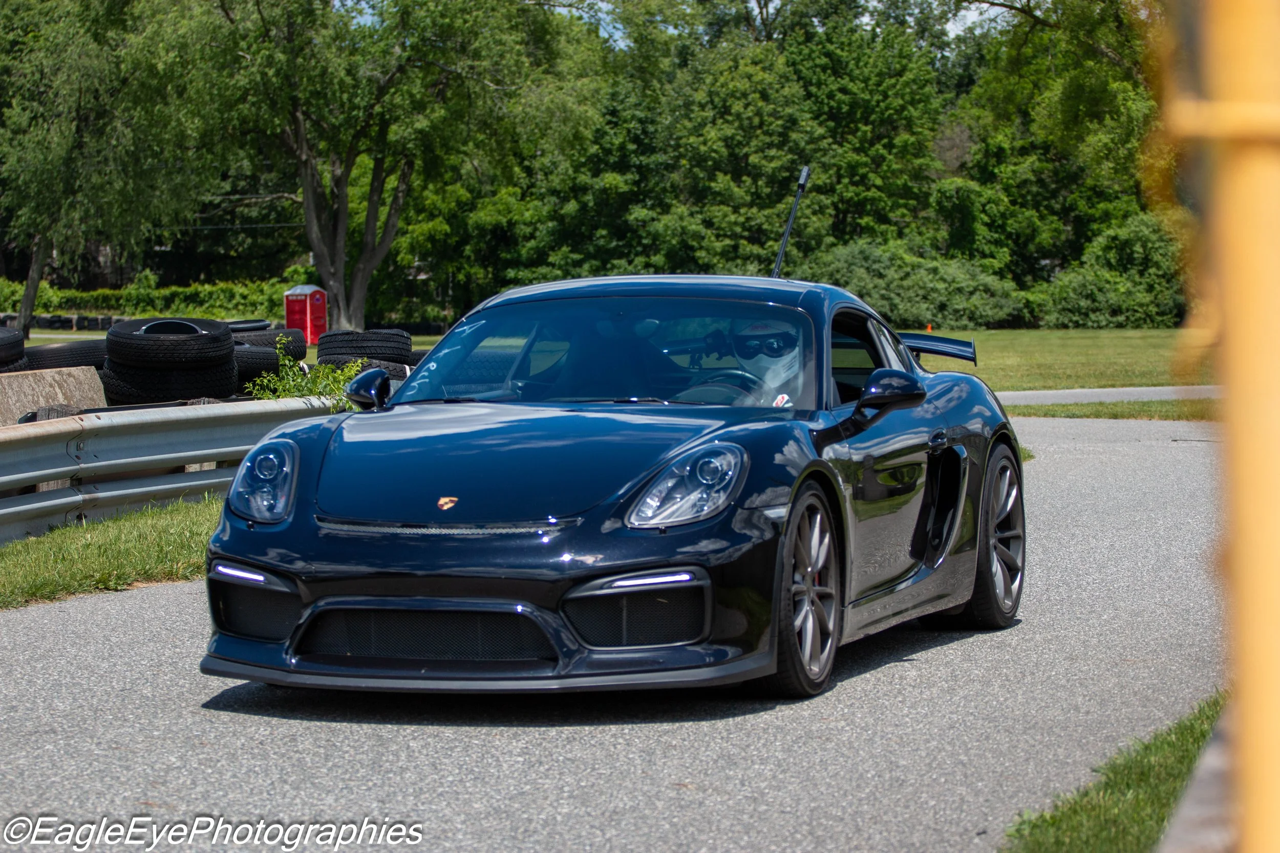 A blue sports car parked on a paved road next to a guardrail with tire stacks and greenery in the background.