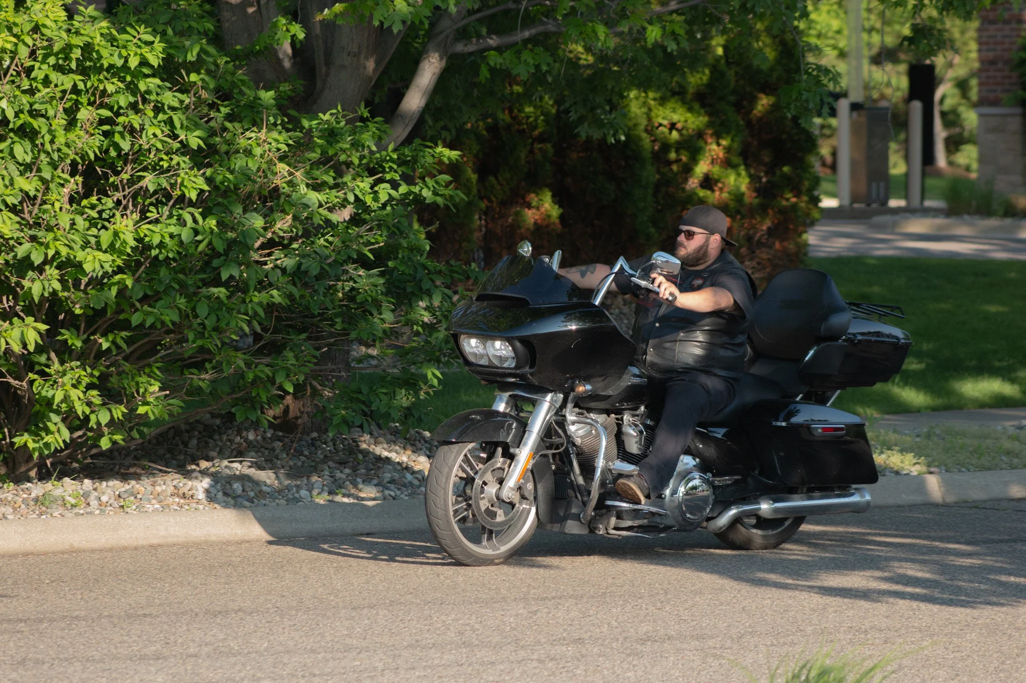 A man wearing sunglasses, a black leather vest, and a backward cap riding a black motorcycle on a tree-lined street.