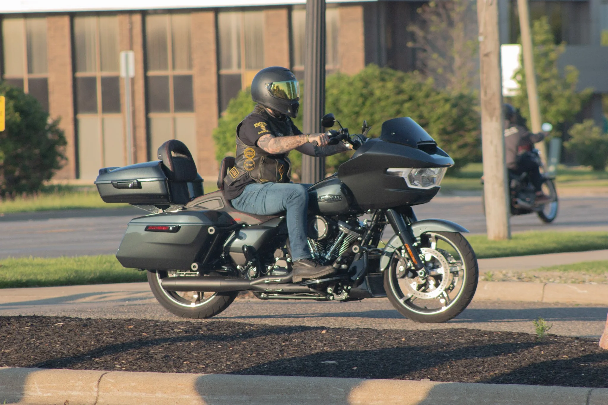 A person riding a black motorcycle with a top box, wearing a black helmet, sunglasses, a vest with patches, and jeans, driving on a street with buildings, trees, and utility poles in the background.