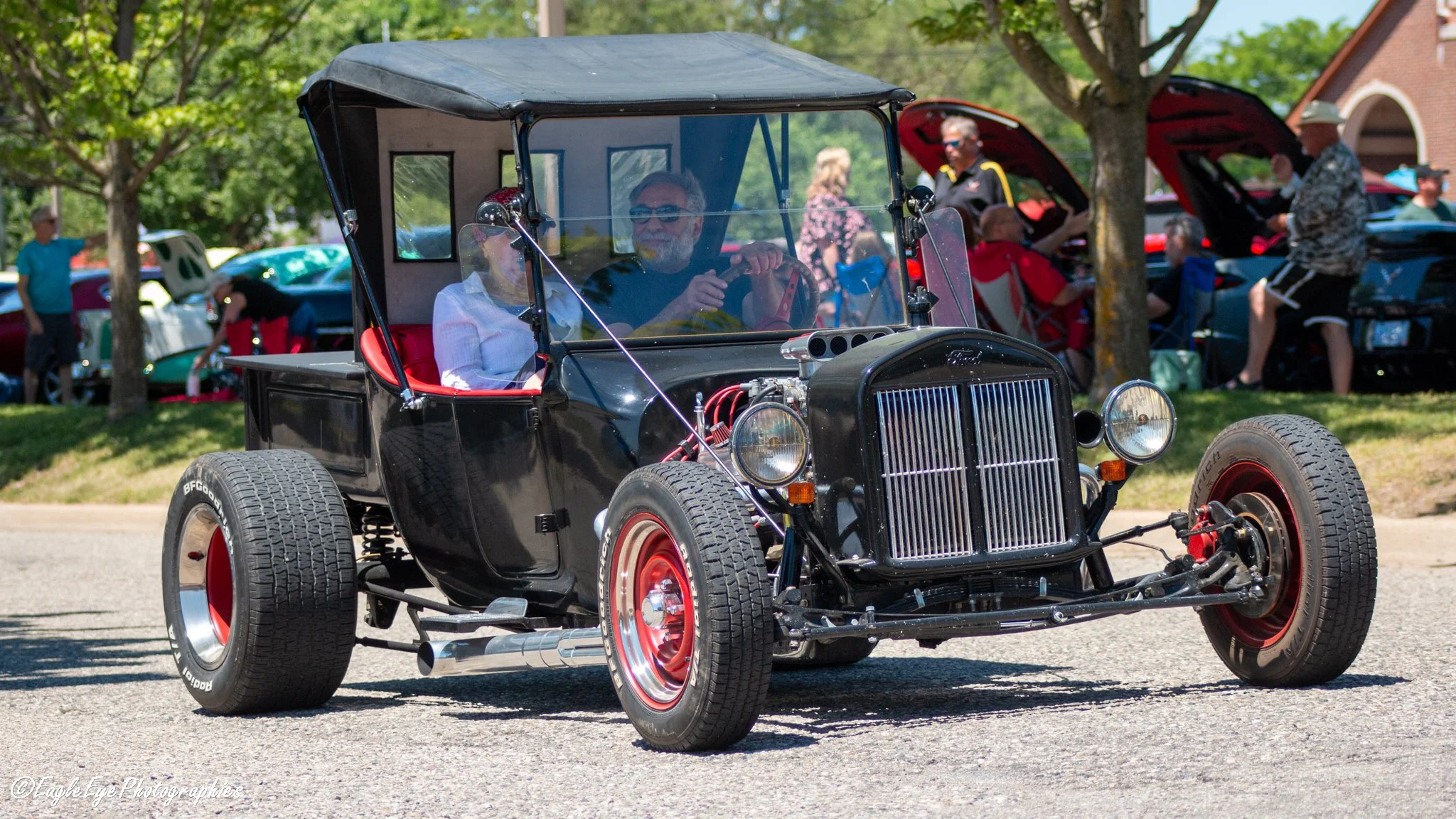A vintage black car with red wheels being driven at an outdoor event surrounded by people and trees.