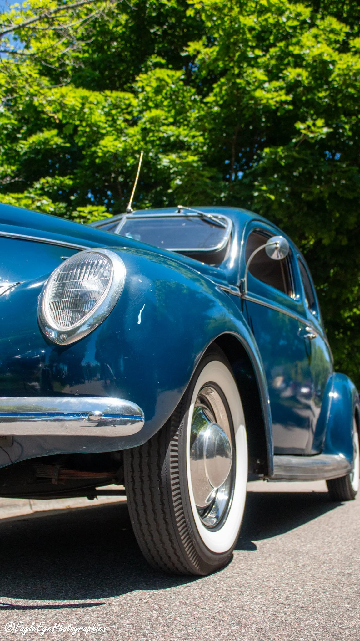 Close-up of a vintage blue car with a white wall tire, headlight, and chrome details, parked on a sunny street with green trees in the background.