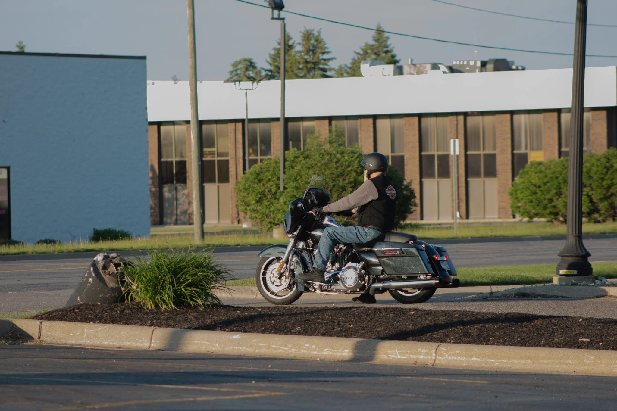 A person riding a black Harley-Davidson motorcycle on a city street, wearing a helmet and jacket, with buildings, trees, and power lines in the background.