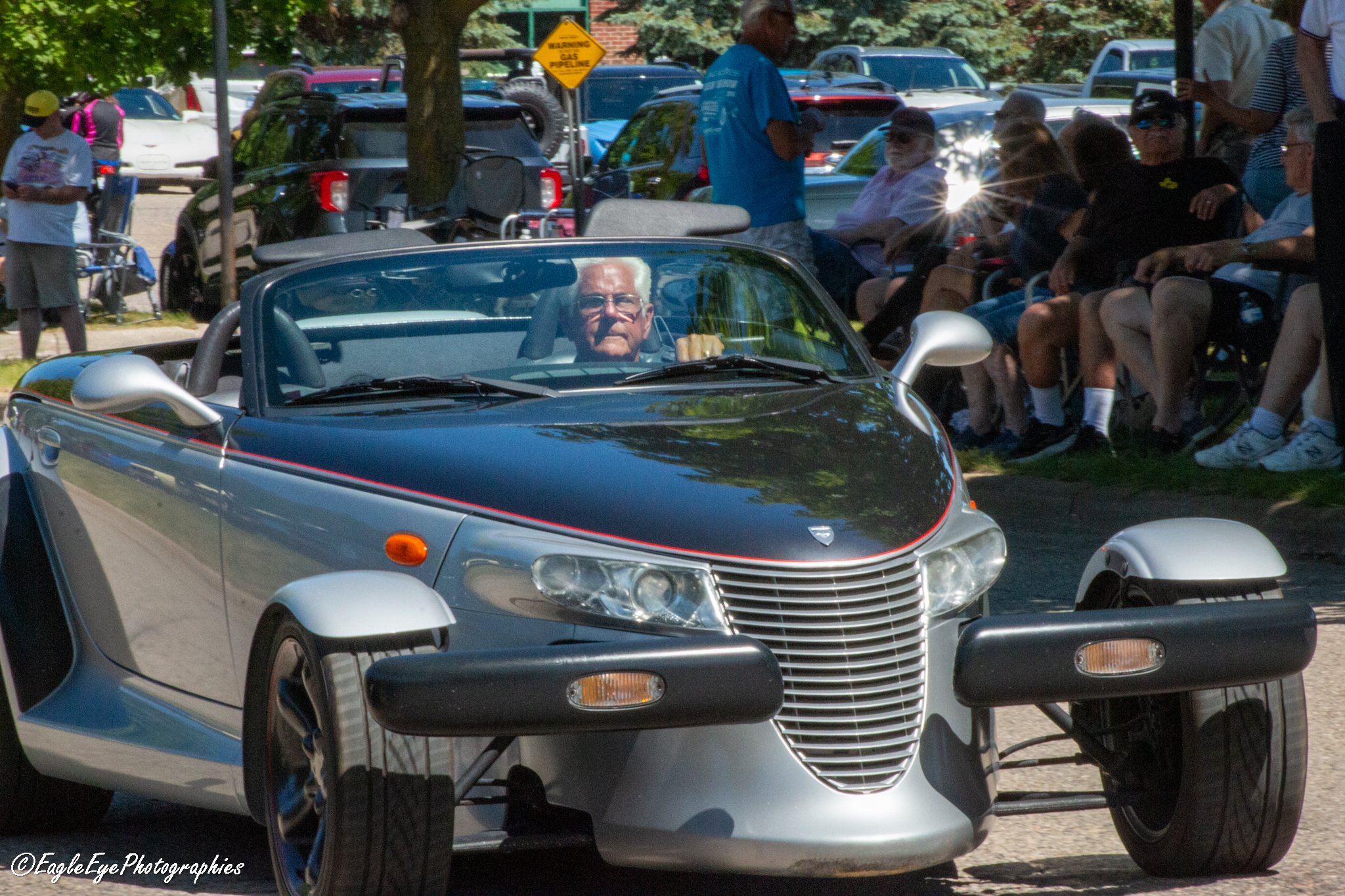 A man with white hair and glasses driving a vintage silver and black Chrysler convertible car, with a crowd of people seated on benches and standing nearby in a park-like setting, trees and parked cars in the background, and a yellow warning sign abo