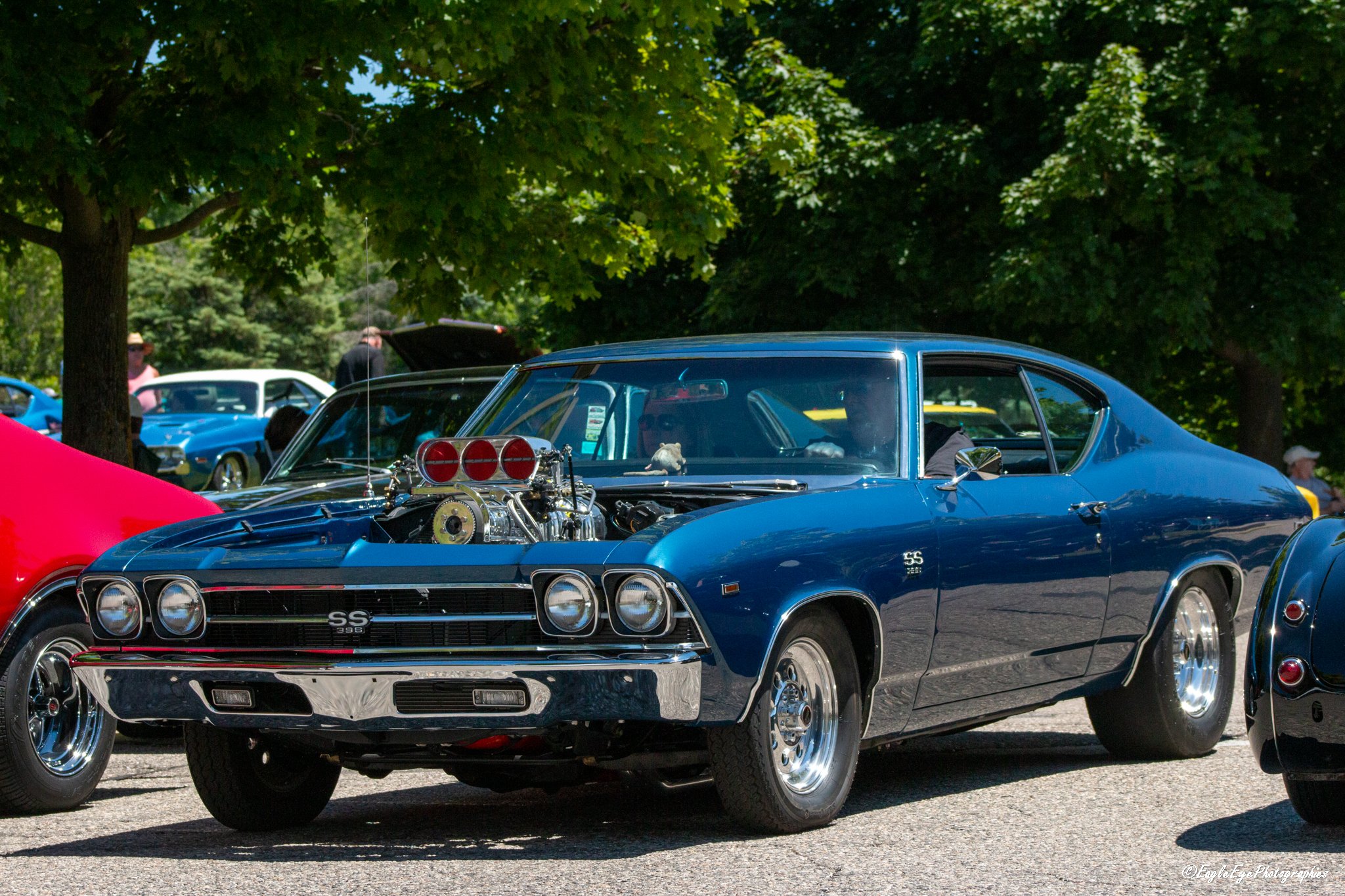 A classic blue Chevrolet muscle car with an exposed engine, parked among other vintage cars at a car show under green trees.