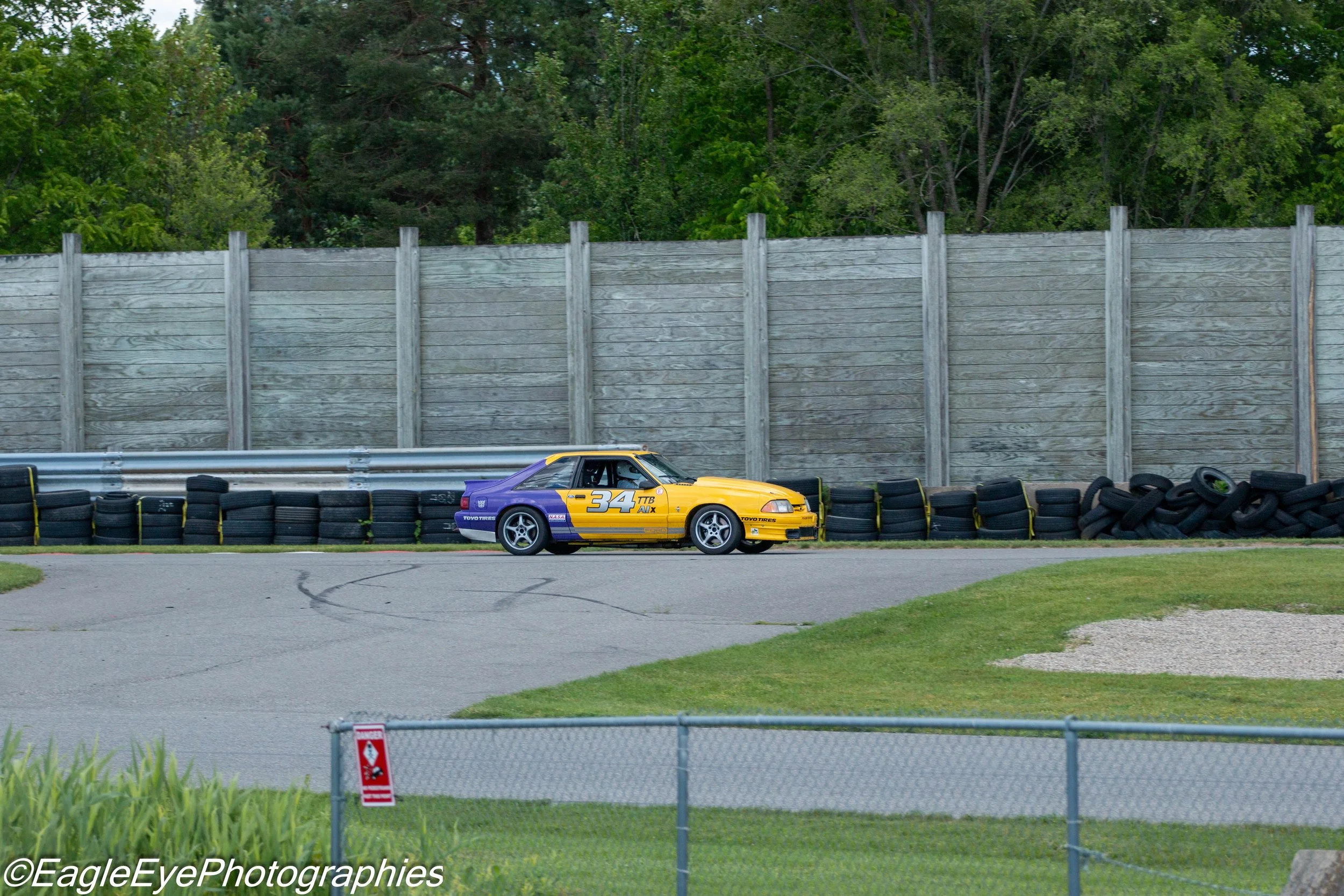 A yellow and purple race car with the number 34 on the side, driving on a racetrack during daytime. It is passing a stack of tires and a fence, with tall green trees and a wooden privacy fence in the background.