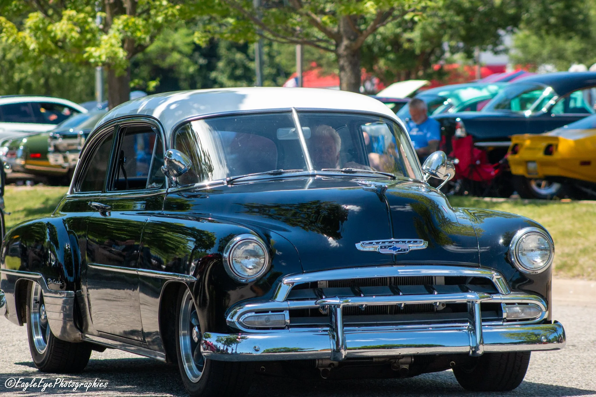 A shiny black vintage Chevrolet car at a car show, with other classic cars in the background and trees providing shade.