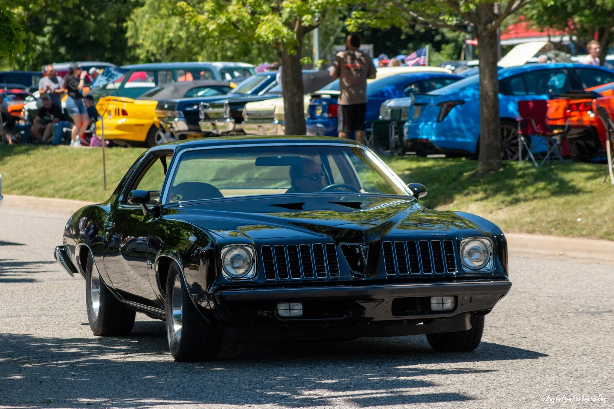 A black vintage Pontiac Firebird car driving at a car show with classic cars parked on grass and people walking and observing in the background.