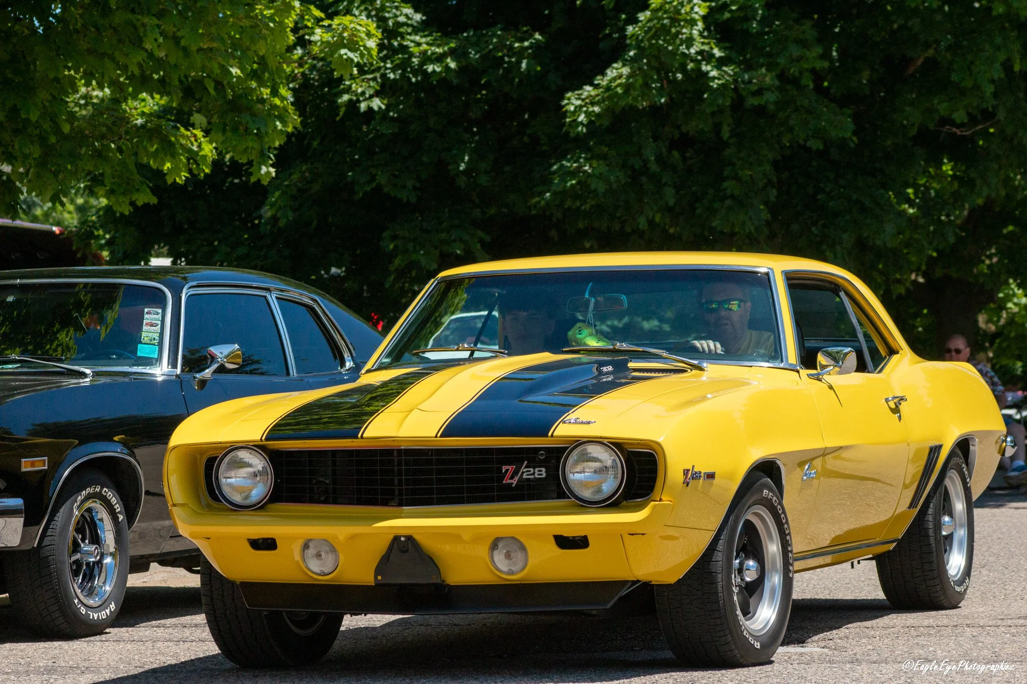 A yellow Chevrolet Z28 muscle car with black racing stripes, parked next to a dark blue car, against a background of green trees on a sunny day.