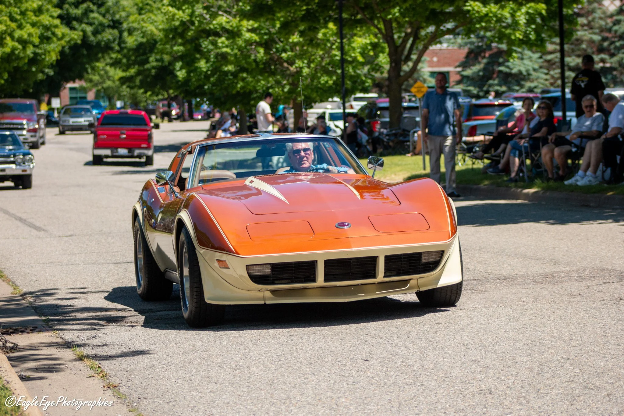 An orange vintage sports car driving down a street during a car show, with people sitting on chairs and trees lining the sidewalk.