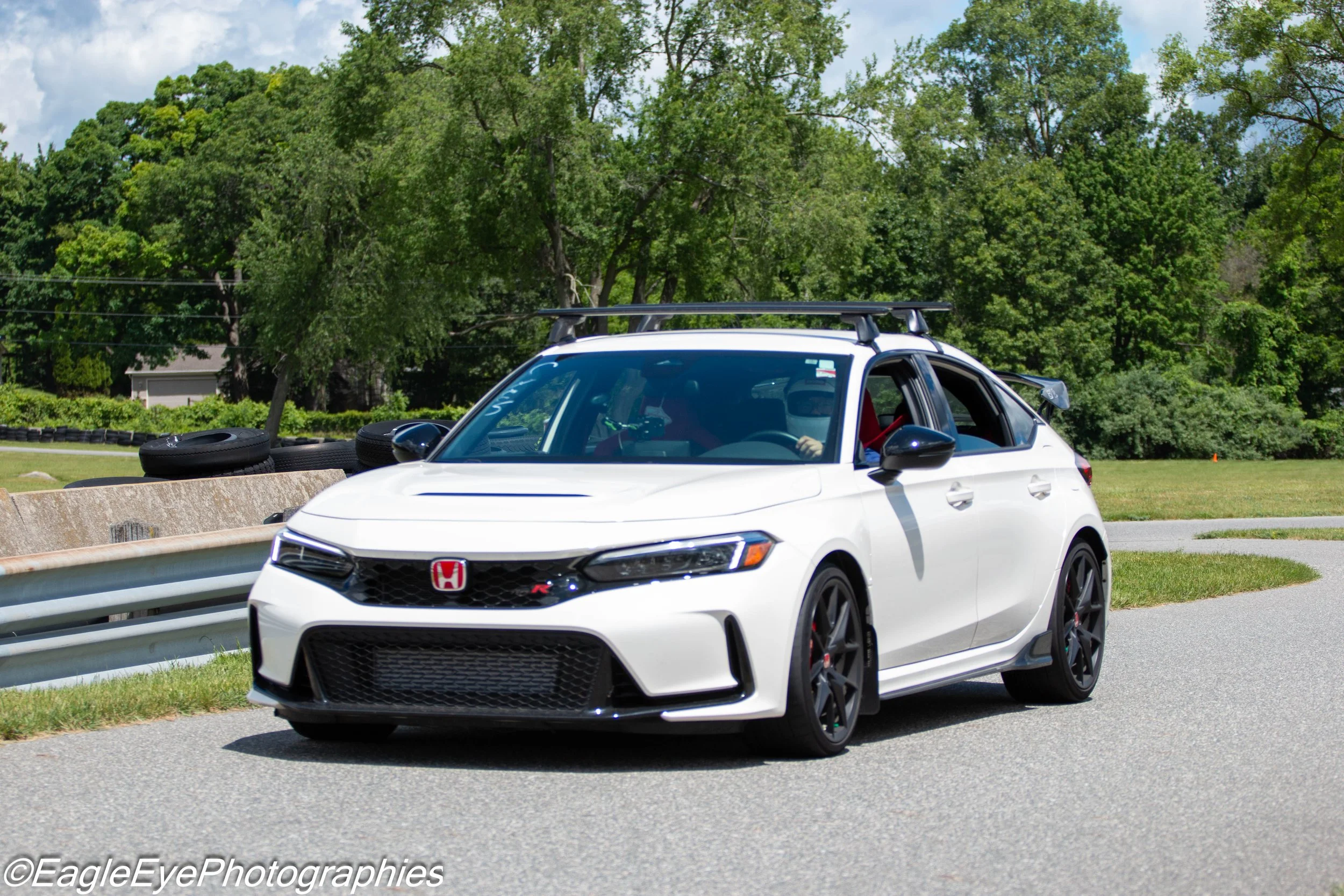 A white Honda Civic Type R with black wheels and a red Honda emblem driving on a paved road. There is a guardrail and green grass on one side, and a background of green leafy trees and blue sky with clouds.