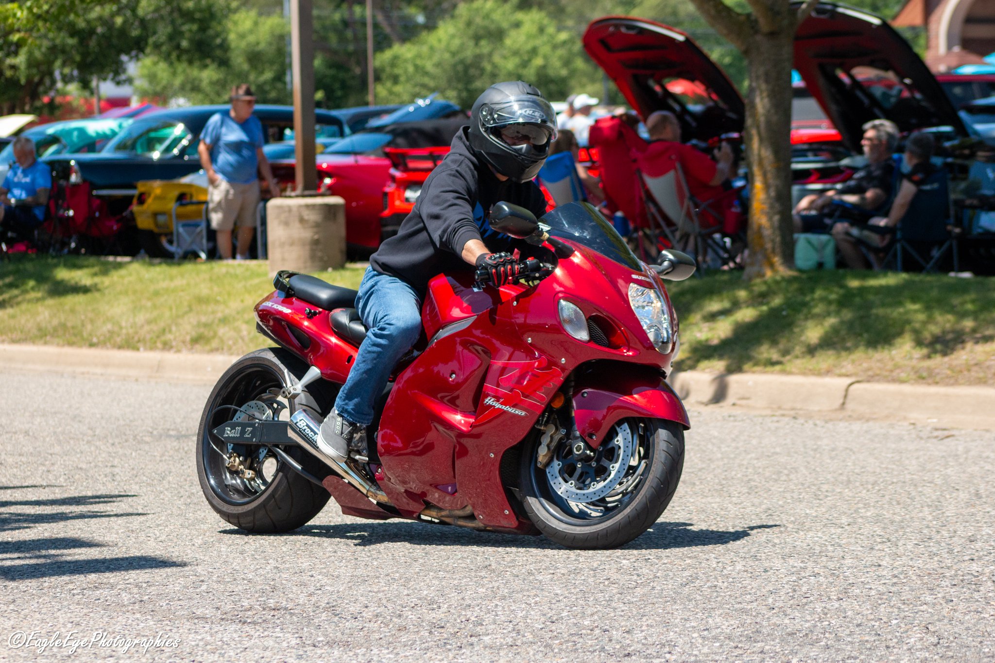 A person wearing a helmet and black hoodie riding a red motorcycle on a street, with a background of cars parked under trees and people near a car show.