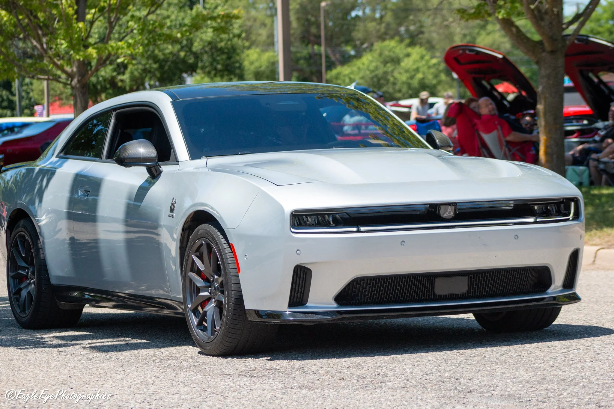 A silver sports car parked outdoors with trees in the background, other cars with open hoods, and people sitting on chairs at a car show or gathering.