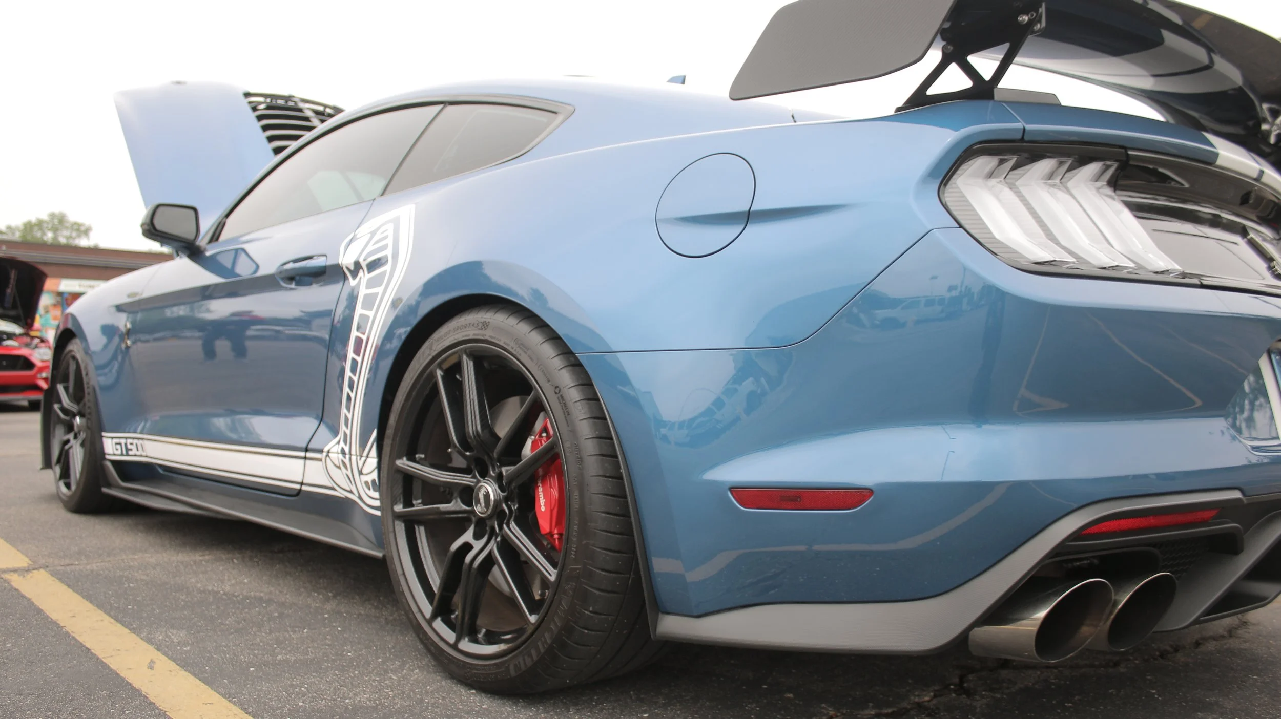 Blue Ford Mustang Shelby GT350 with black wheels and red brake calipers, parked at a car show with other cars in the background.