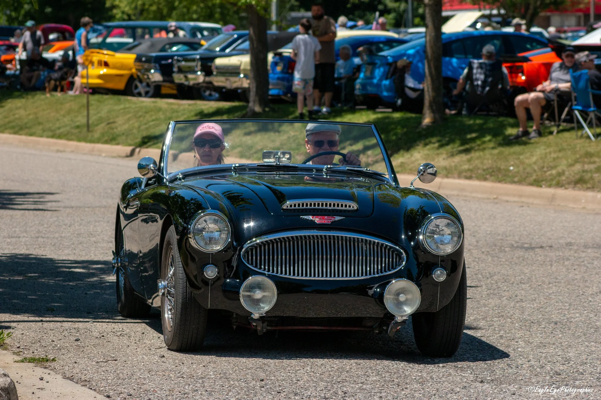 A black vintage convertible sports car driving on a road with two people inside wearing sunglasses and hats. In the background, there are people, parked cars, and trees at an outdoor car show or event.