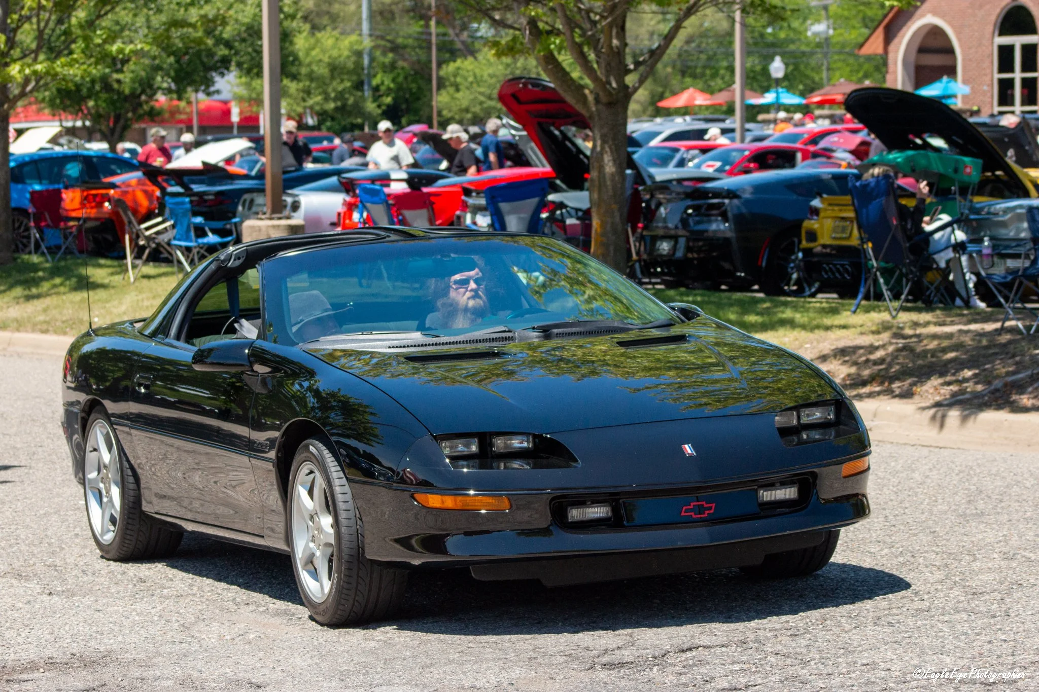 Black Chevrolet sports car parked outdoors at a car show with its hood closed, while a crowd and multiple other cars are visible in the background under trees and on a sunny day.