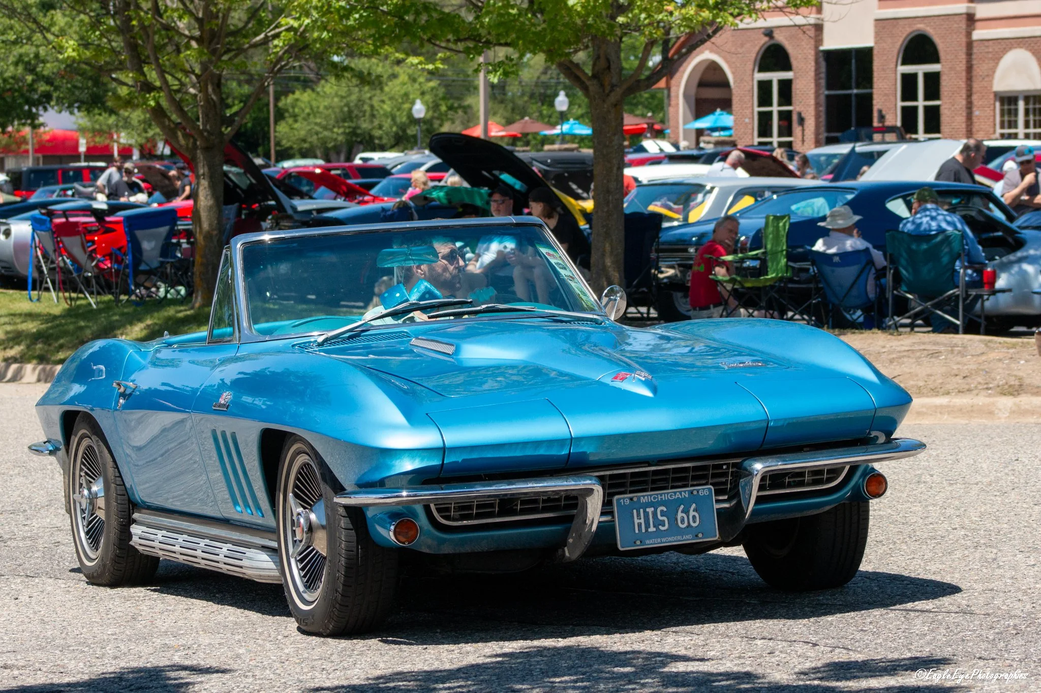 A classic blue Chevrolet Corvette with Michigan license plate 'HIS 66' parked at a car show with other cars and people in the background.