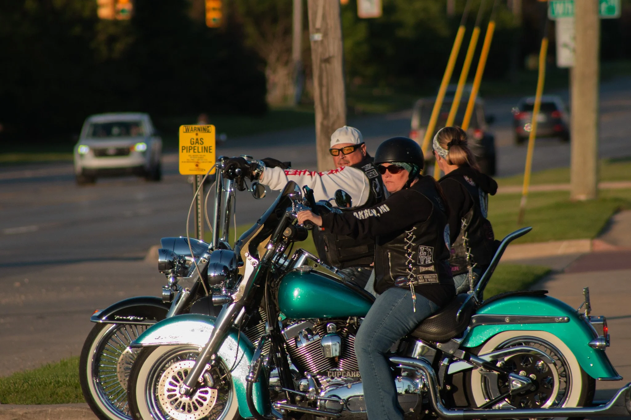Group of people riding motorcycles on a street during daylight with a warning sign about a gas pipeline in the background.