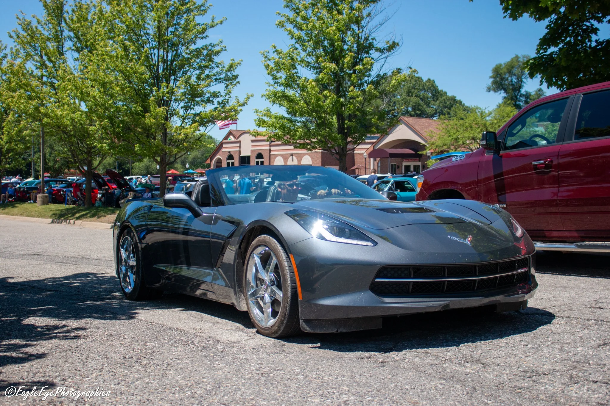 A shiny black Chevrolet Corvette convertible with a driver inside, parked in a busy parking lot on a sunny day. There are green trees and a brick building with an American flag in the background.