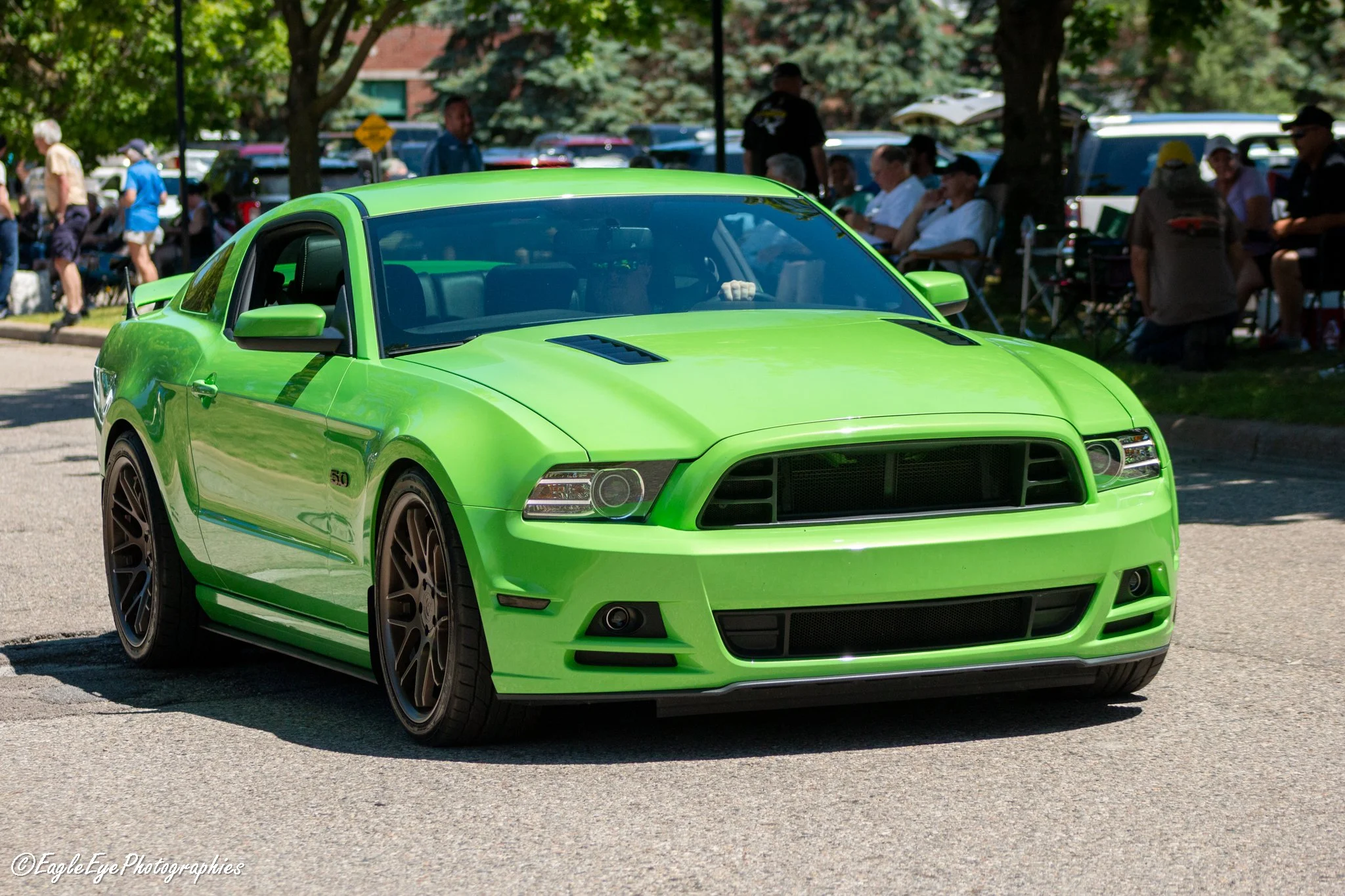 A bright green sports car with black tires on a street, with people sitting and walking in the background shaded by trees.