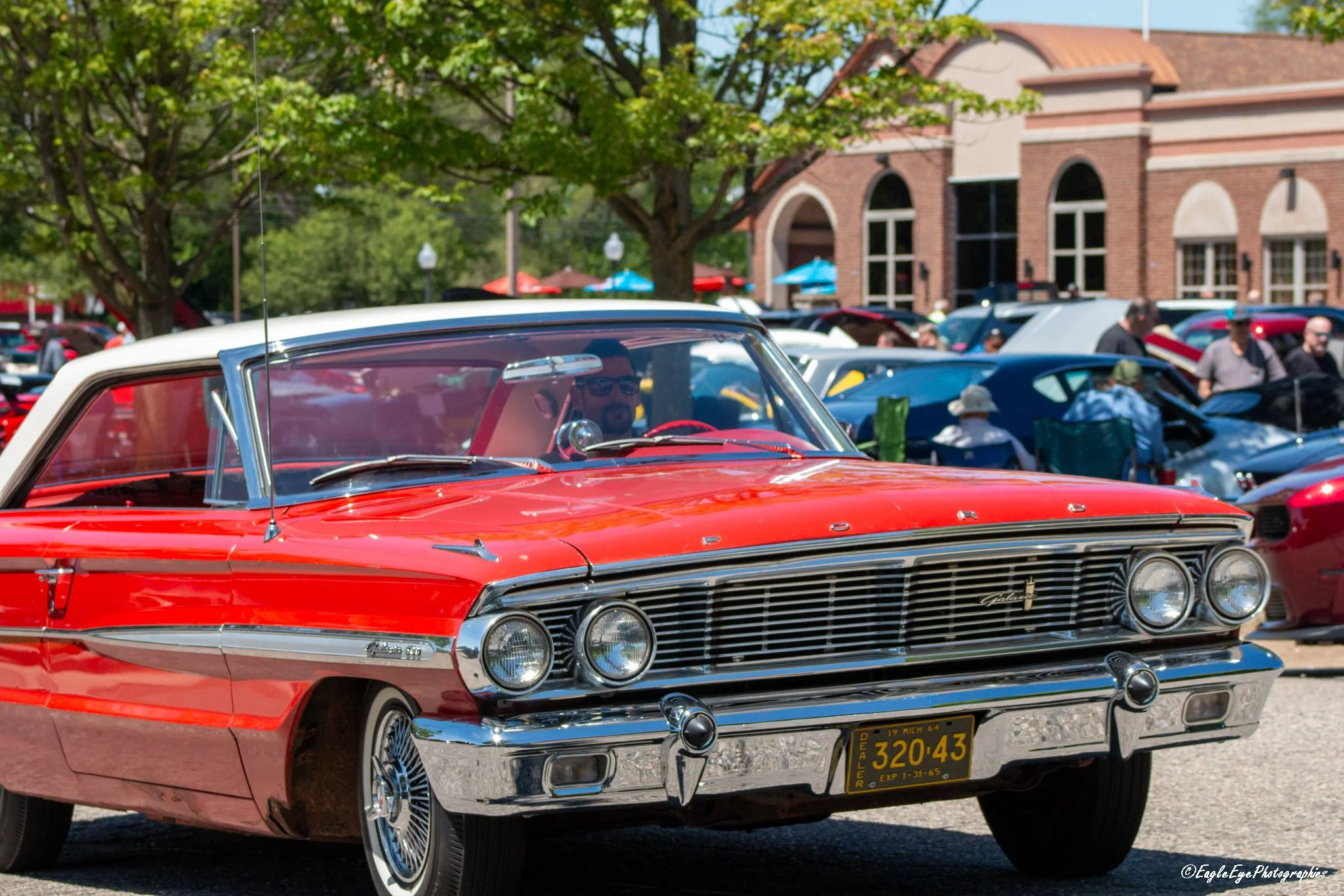 A close-up view of a red vintage Ford Galaxie car parked outdoors during daytime at a classic car show, with other cars and people in the background, trees, and a brick building.