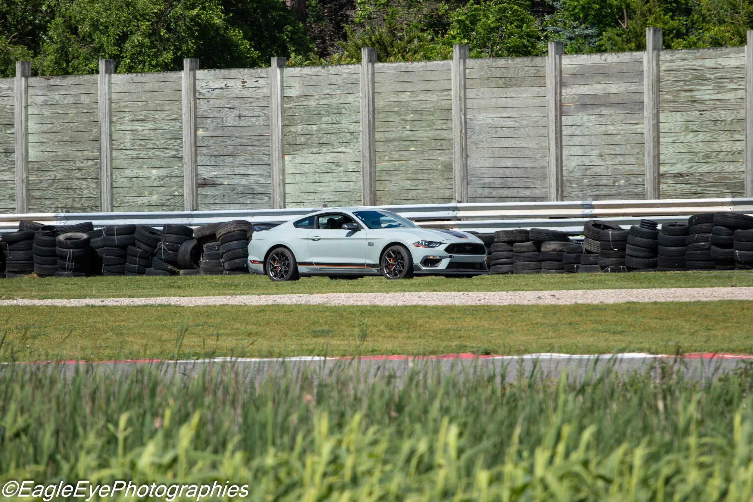 A white sports car with black racing stripes parked behind a row of stacked tires at a race track, with a tall wooden sound barrier and green trees in the background.