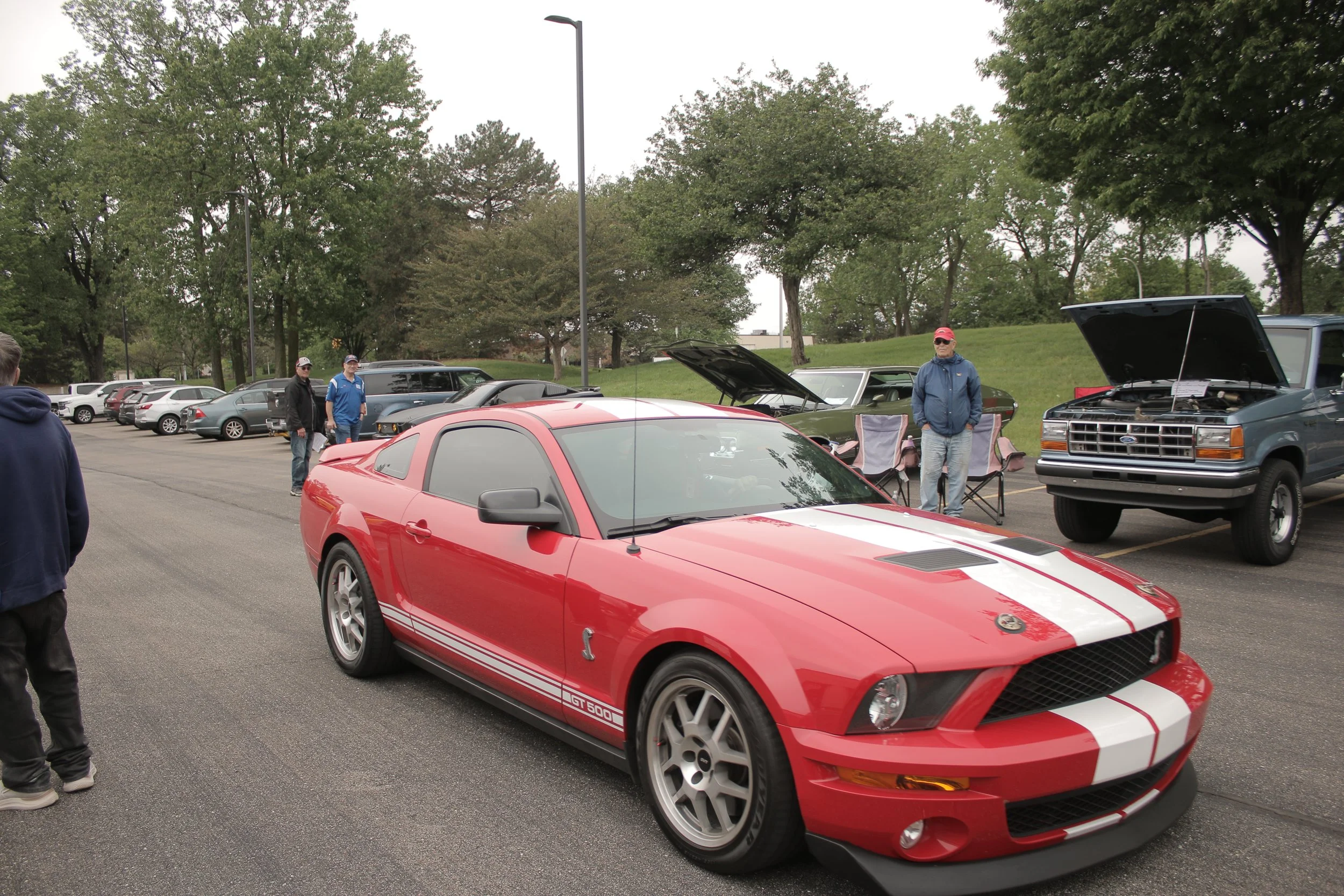 A parking lot with several cars, including a red sports car in the foreground and mostly older vehicles, with trees and lampposts in the background. There are a few people walking and standing near the cars, some wearing casual clothing.