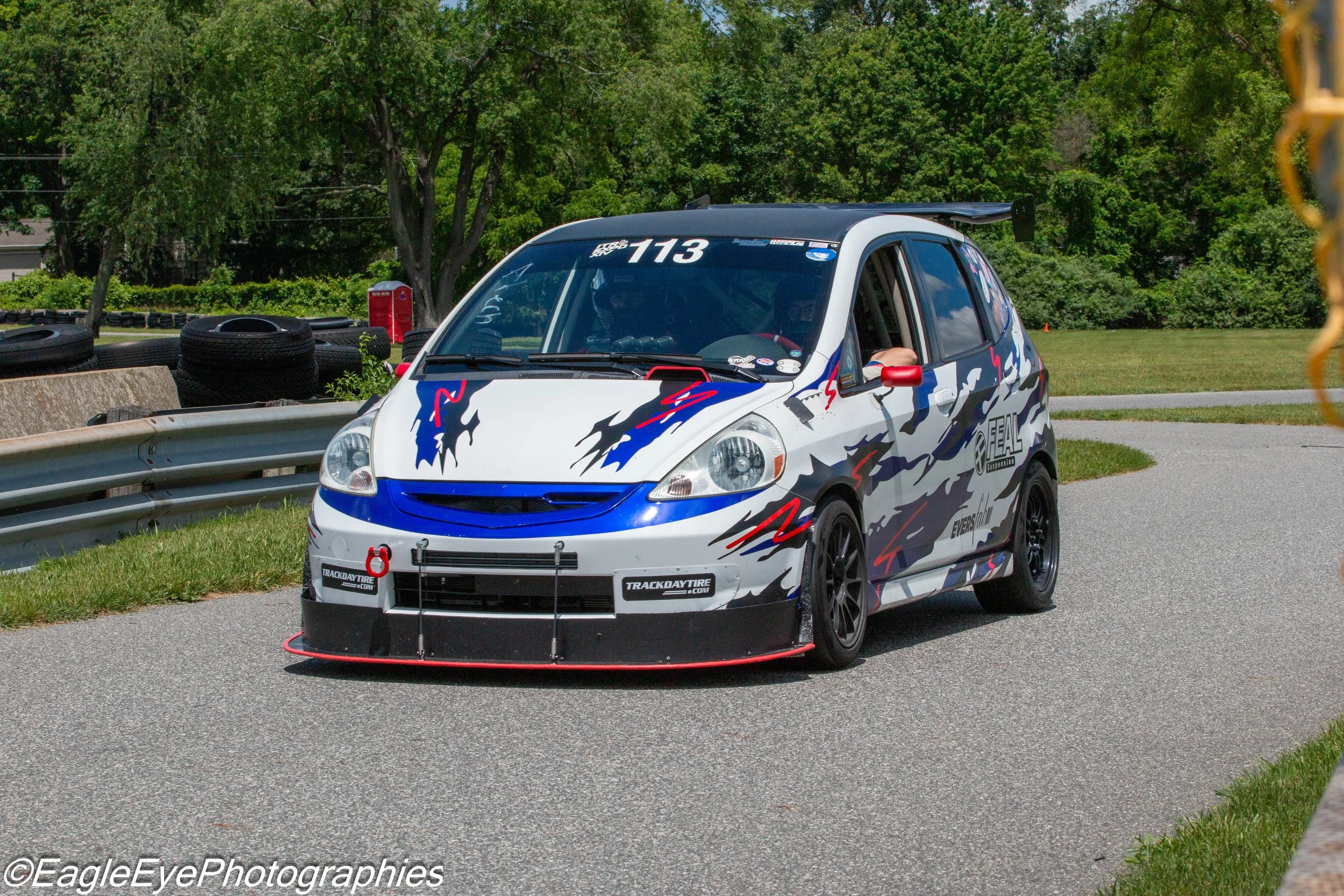 A small white race car with blue and red graphics, the number 113 on the windshield, and sponsors' logos parked on a paved area next to a grassy field with tires and trees in the background.
