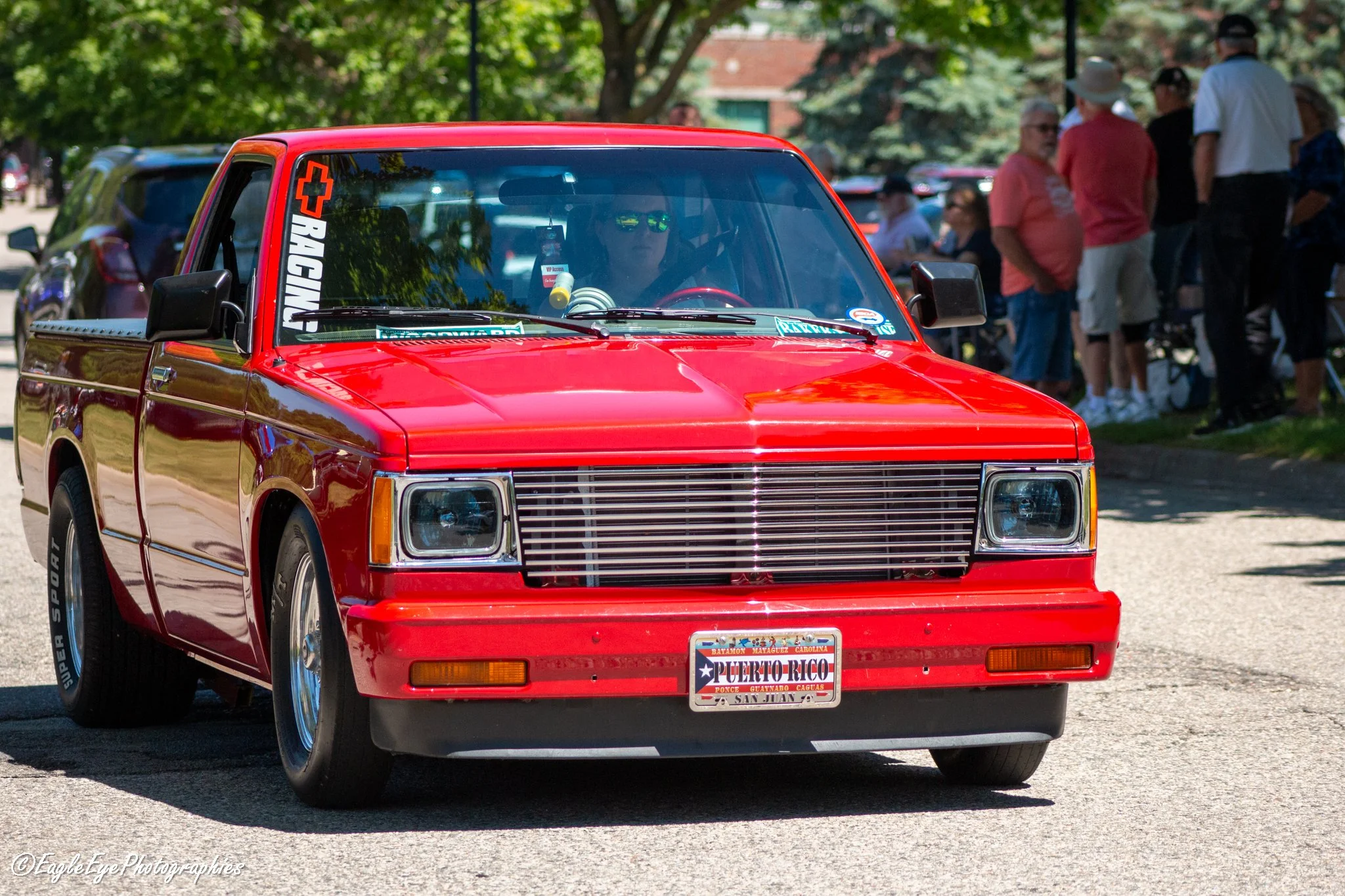 Red pickup truck with a Puerto Rican license plate driving on a street during a parade or gathering with people standing in the background under trees.