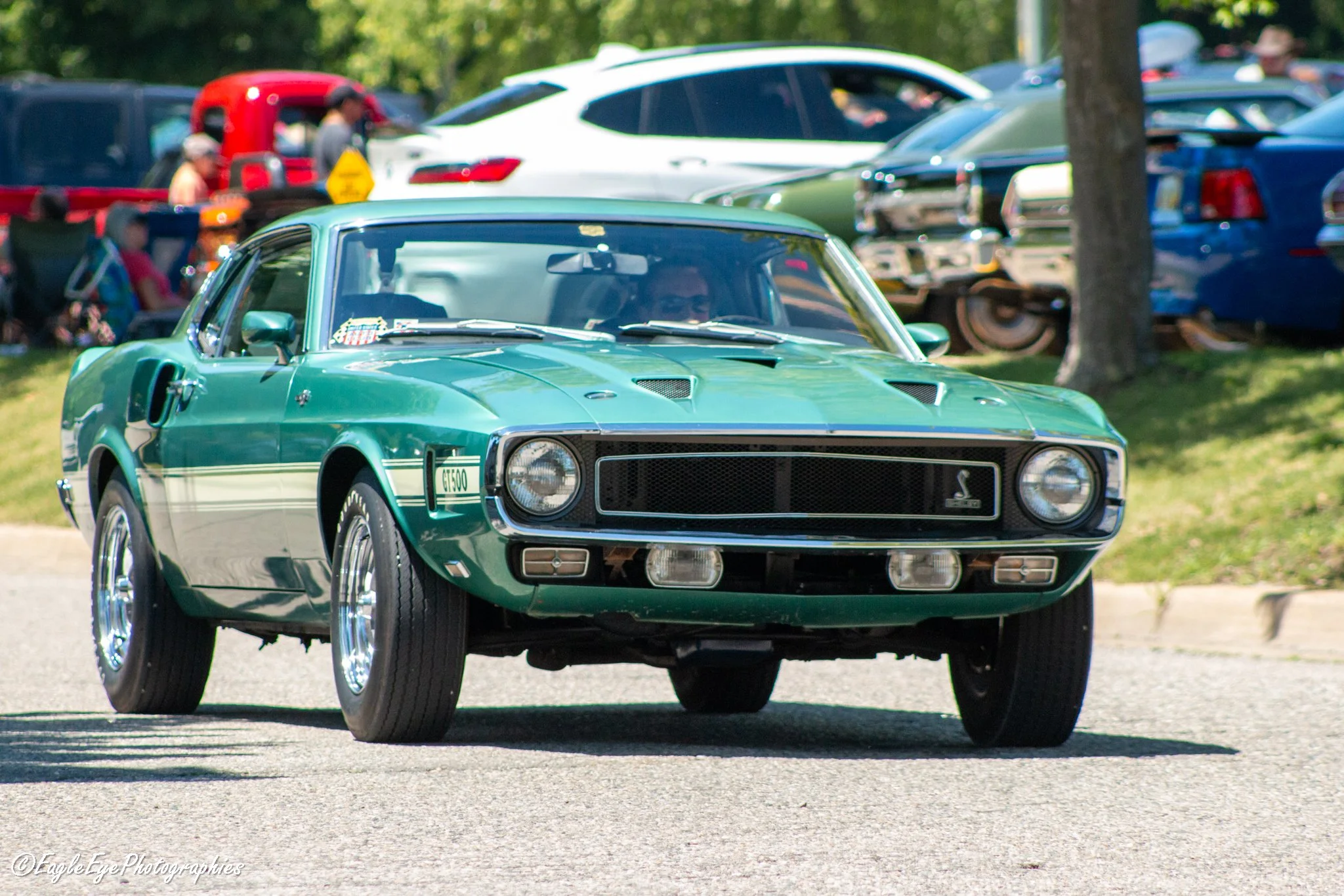 A green vintage muscle car driving on a road with a car show or gathering in the background, featuring tents and parked cars, at a sunny outdoor event.