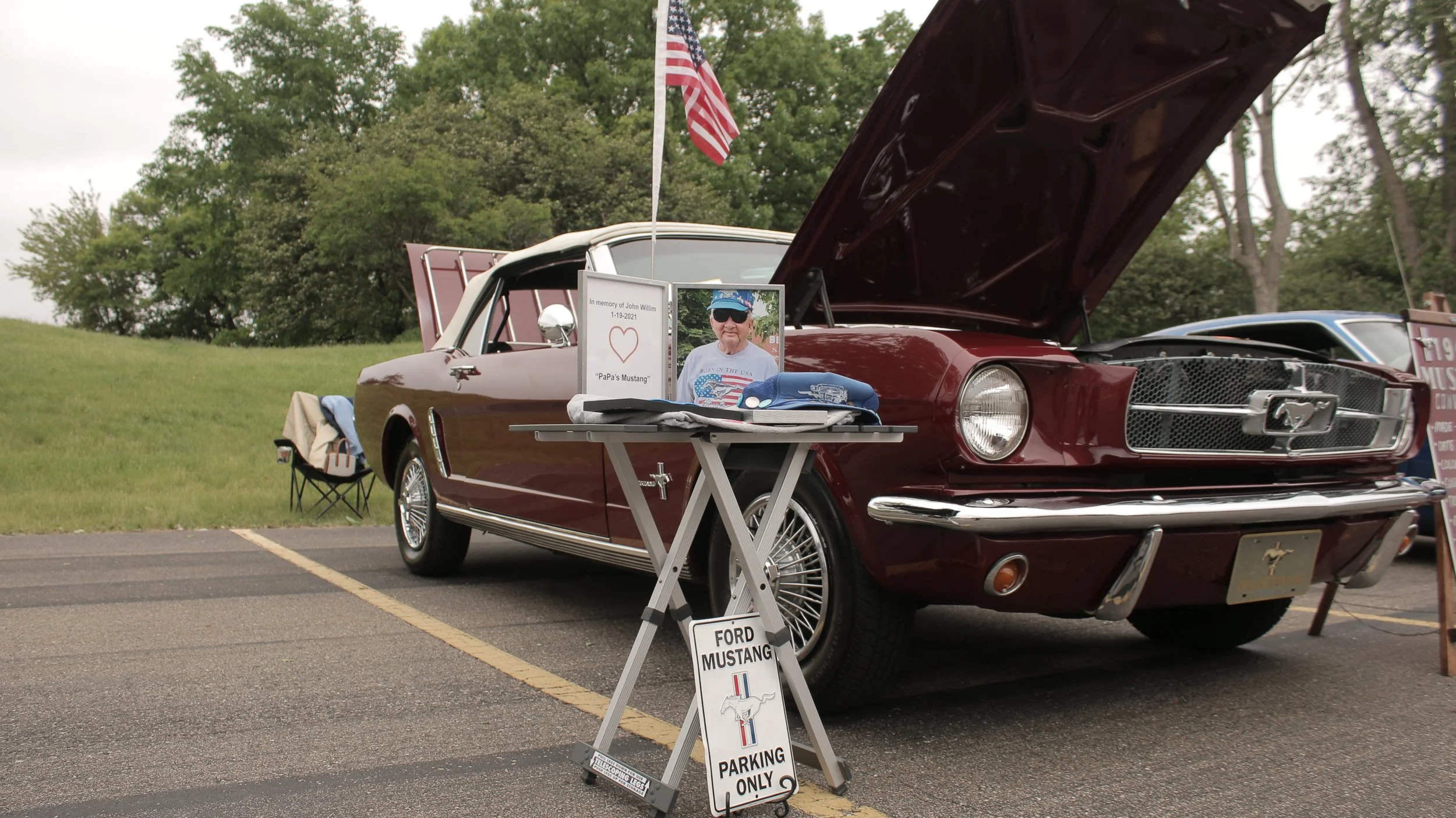 A vintage red Ford Mustang with its hood open, parked in a lot with a grassy area and trees in the background. There is a small table in front displaying items and a framed photo of a man with a sign memorializing John Willim. An American flag is mou